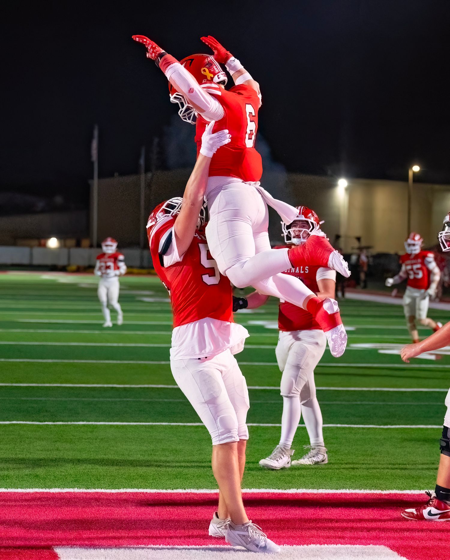Football players celebrate with a victory lift on the field during a nighttime game.