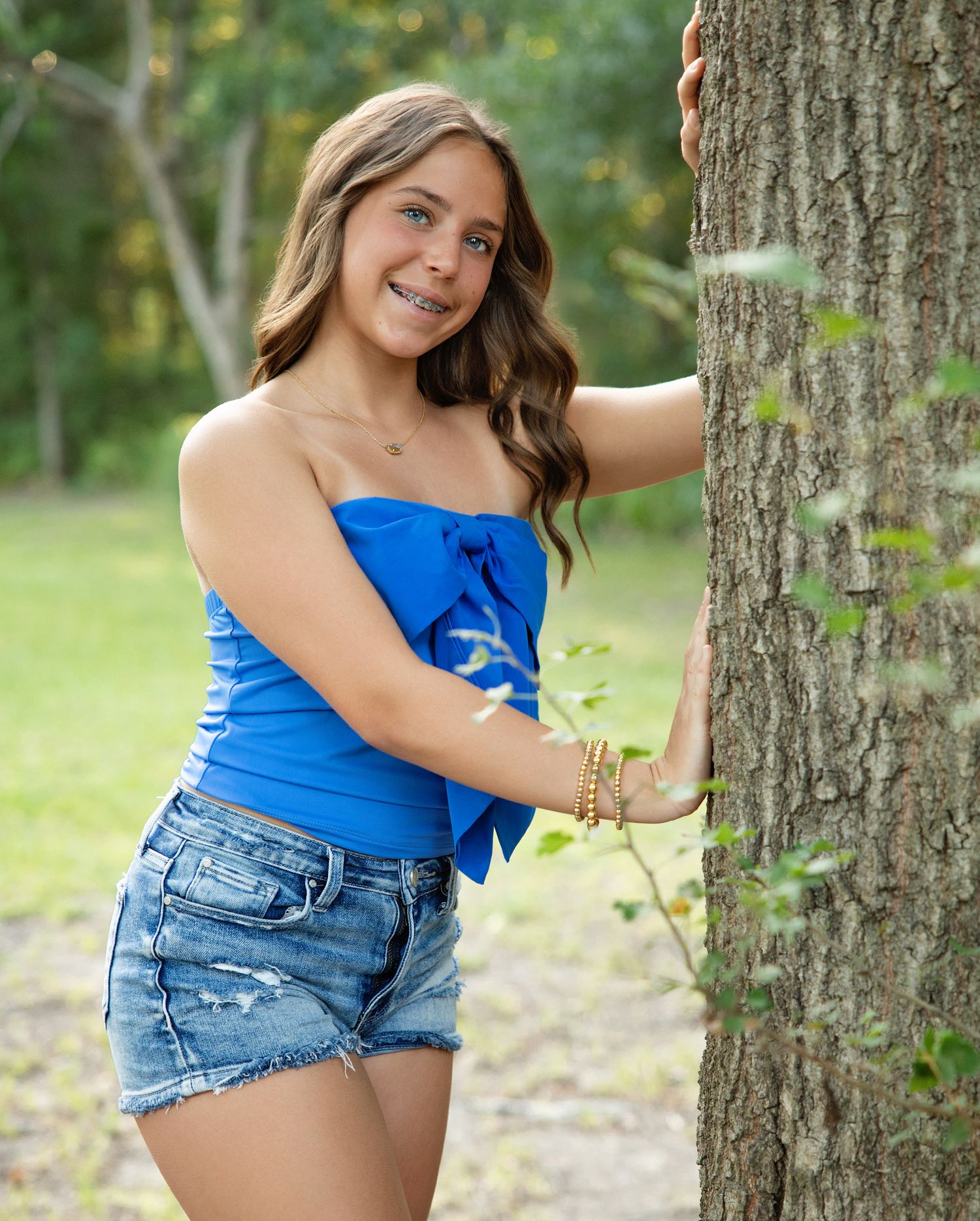 Summer portrait session in nature with blue top and denim shorts against tree backdrop.