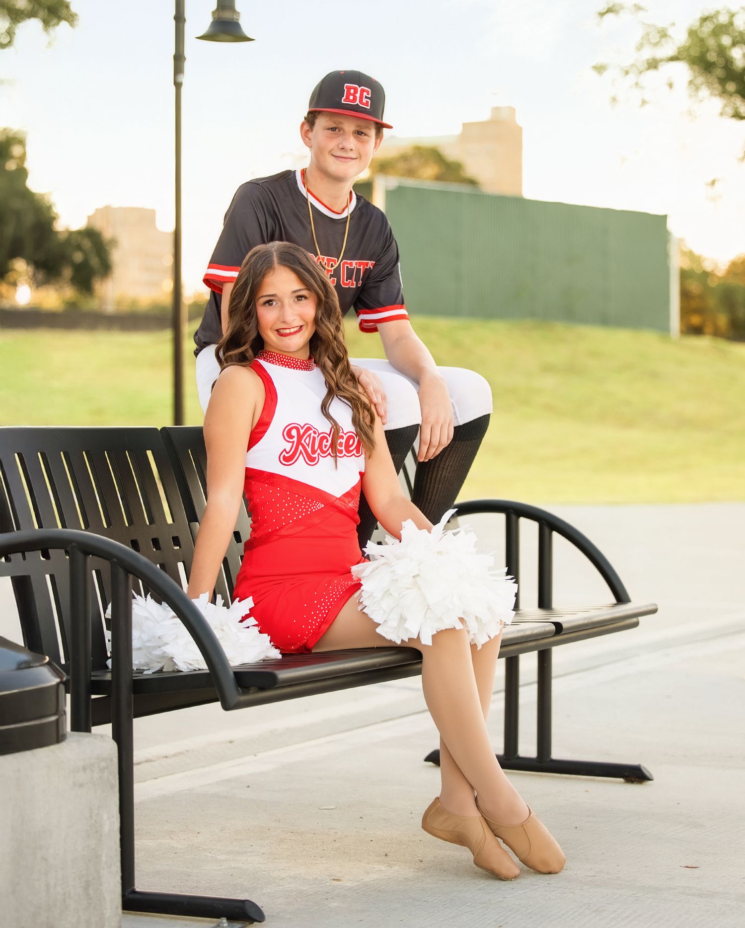 A cheerleader in red and white uniform sits on a black bench outdoors during a sunny day.