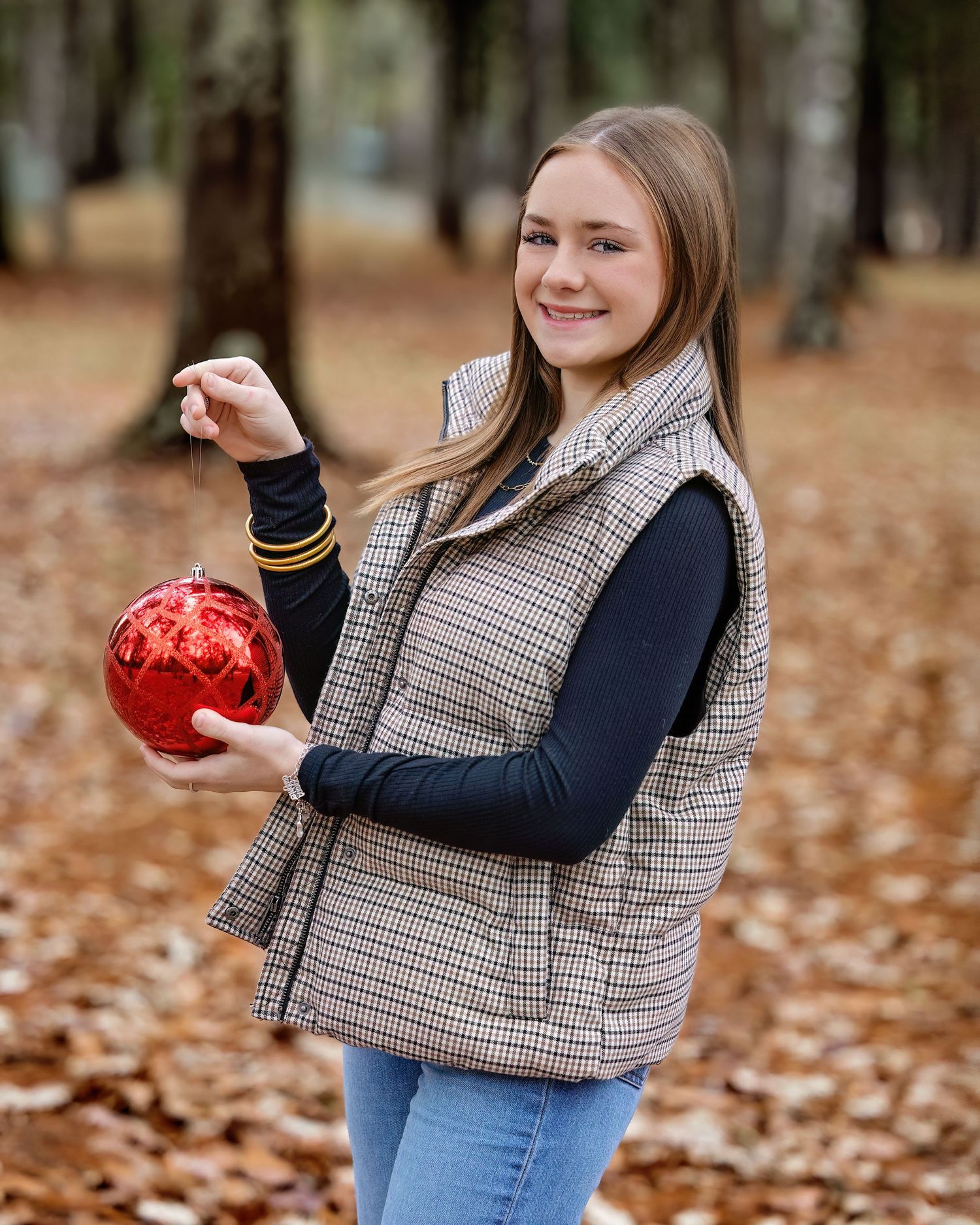 Person in plaid vest and blue jeans holds red ornament while posing in autumn leaves at outdoor photoshoot.