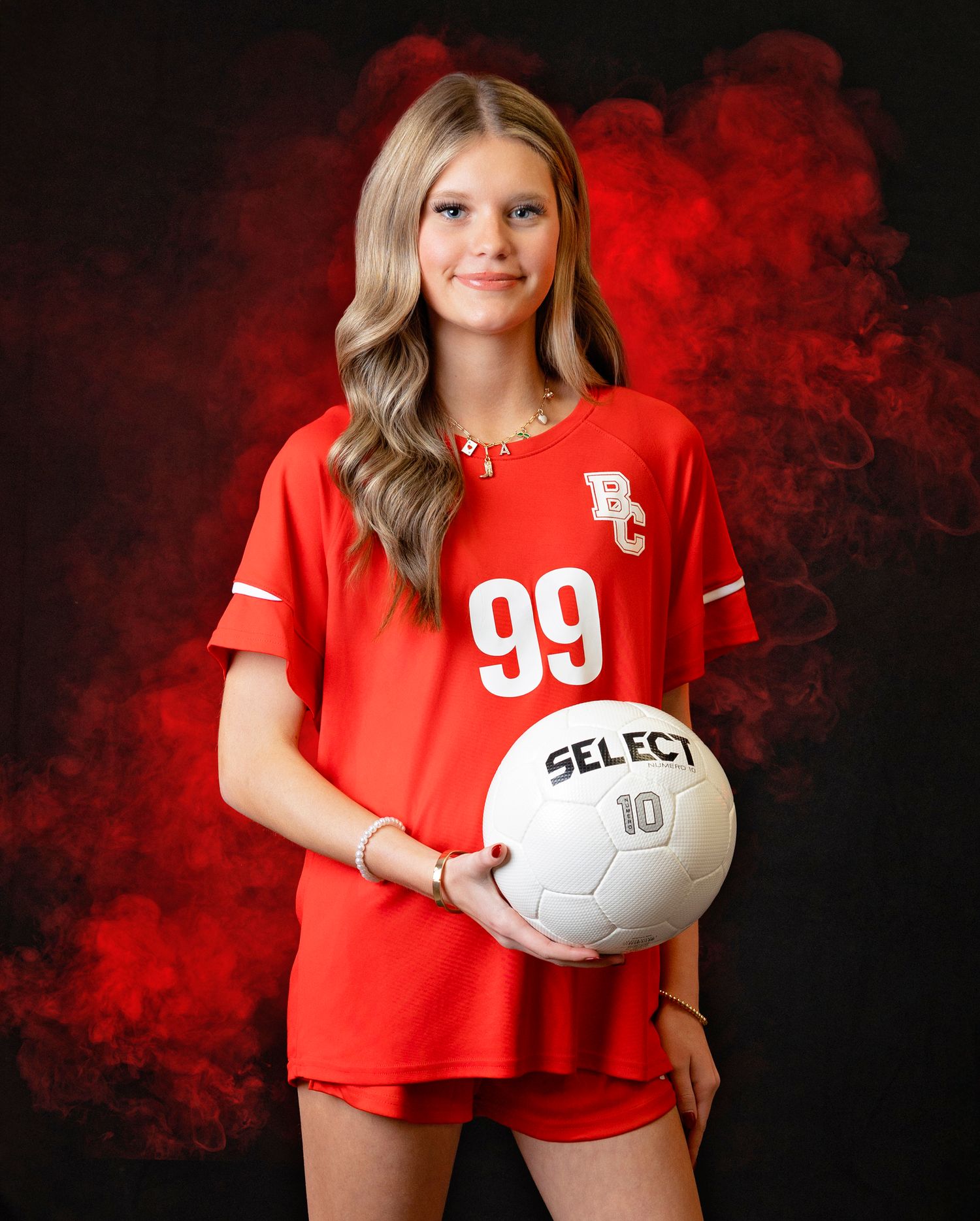 Soccer player in red jersey number 99 poses with white Select ball against dramatic red smoke background.