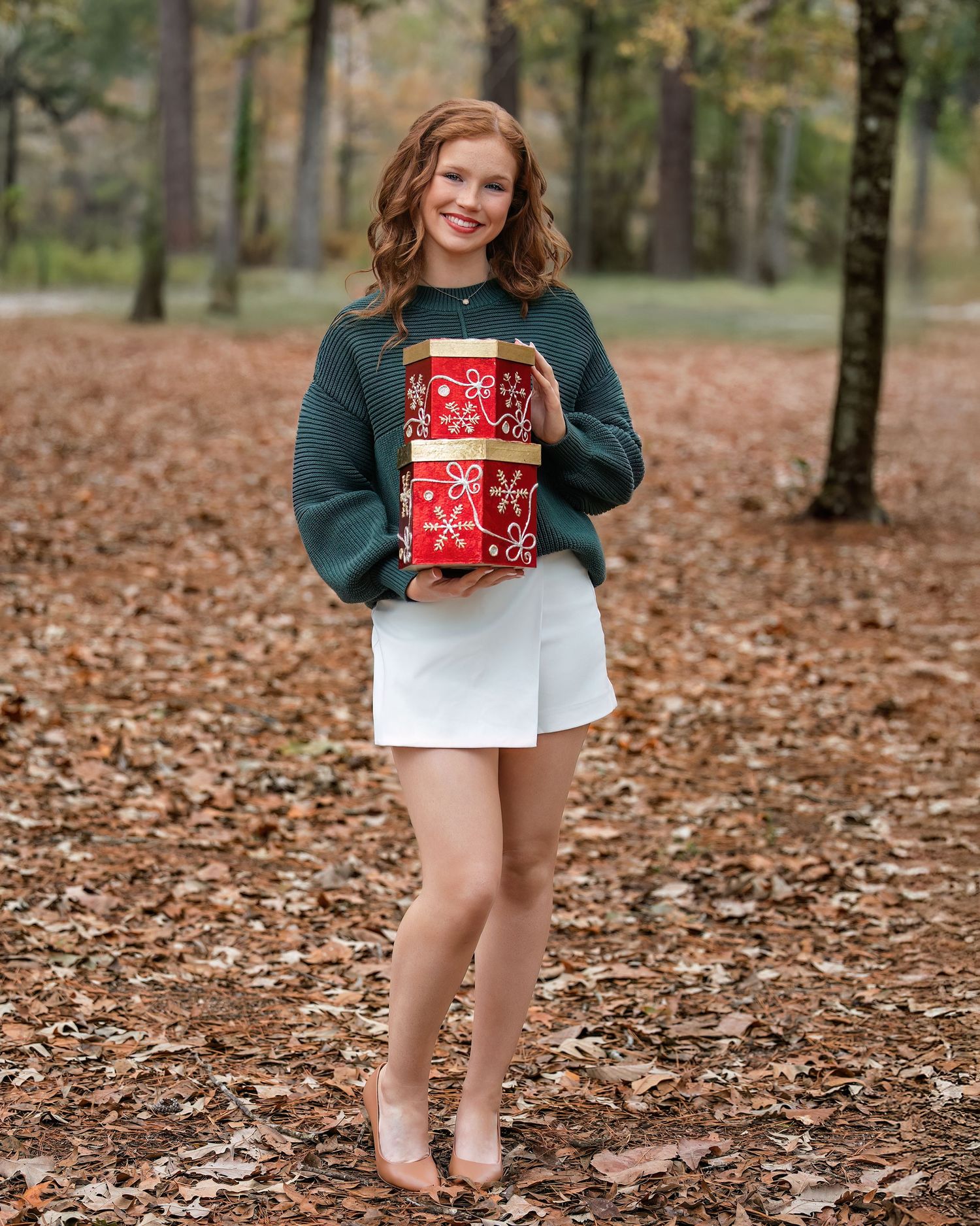 Vidor High School Senior wearing a teal sweater and white skirt holds wrapped Christmas presents while standing on autumn leaves.