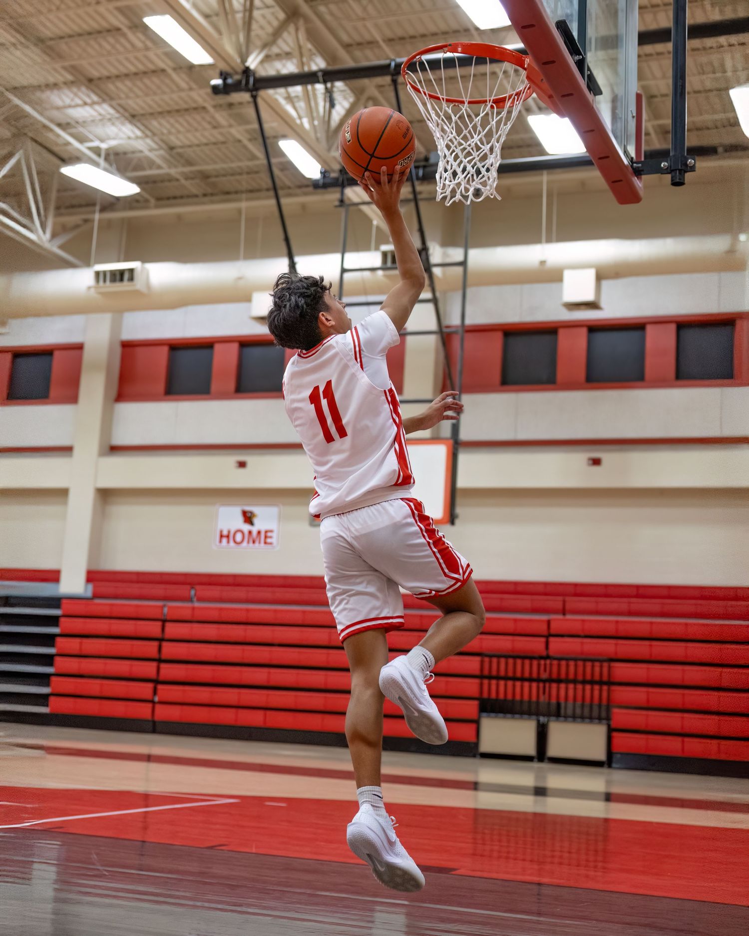 Basketball player in white jersey performs a layup sequence in an indoor gymnasium with red bleachers.