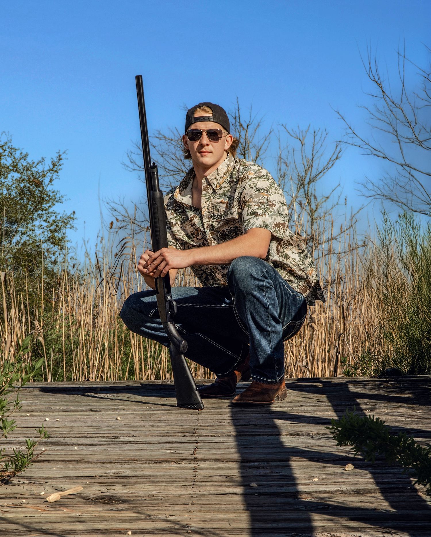 Young man in camo shirt and sunglasses crouching on wooden dock holding rifle outdoors.