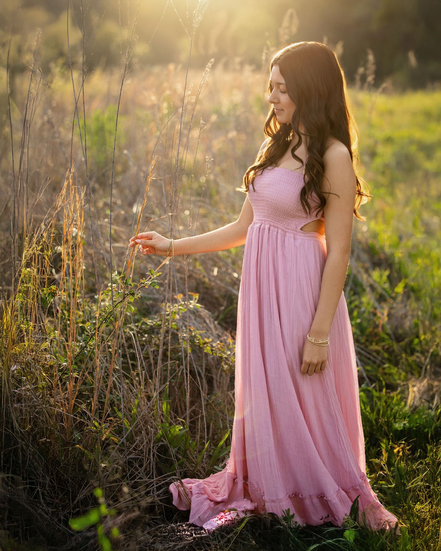 Young woman in pink maxi dress touching wildflowers in golden sunlit field at sunset.
