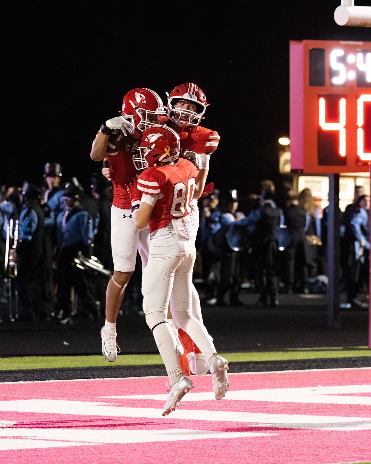 Football player in red uniform leaps into the end zone to score a touchdown during a night game.