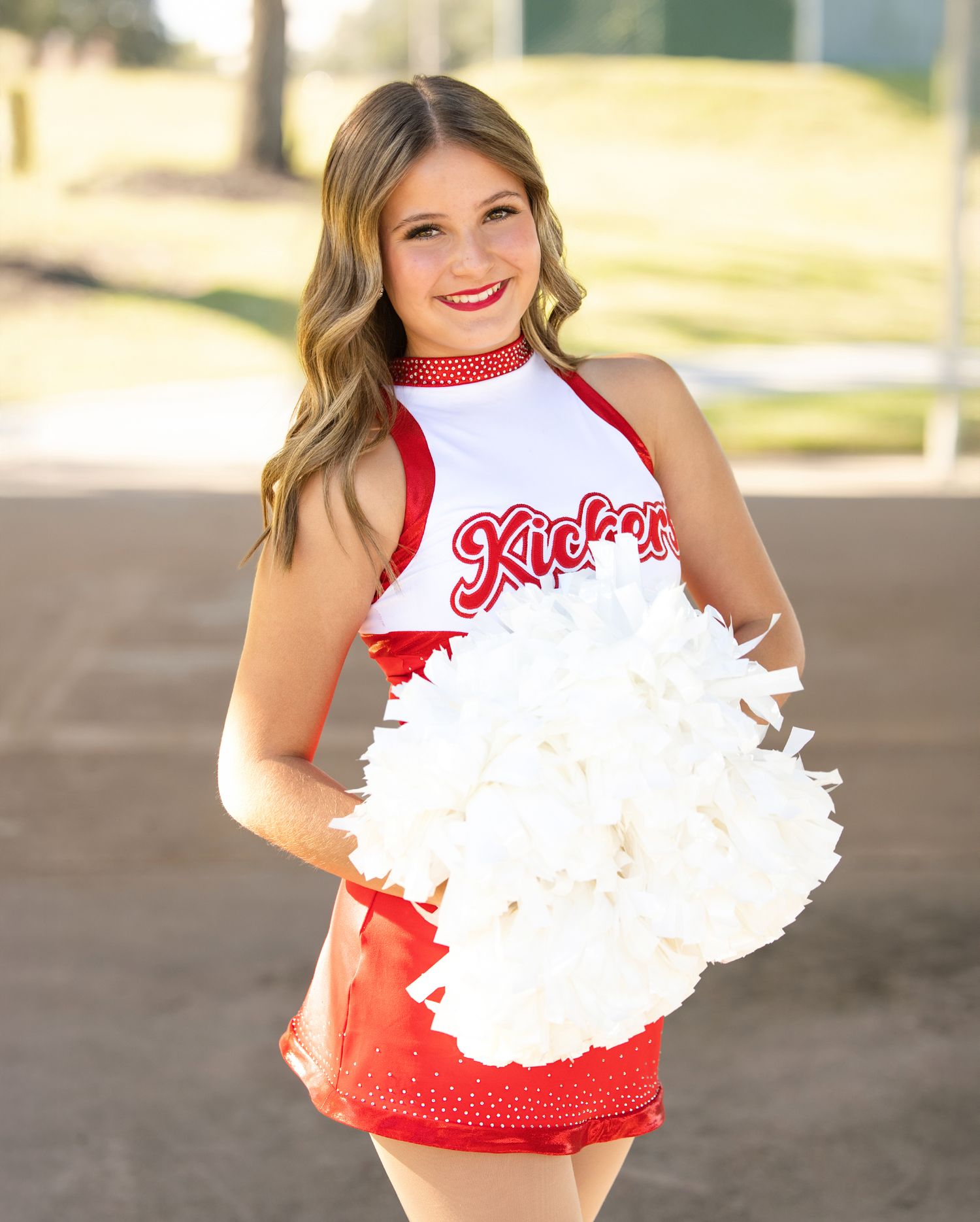 Cheerleader wearing red and white uniform performs with white pom poms outdoors.