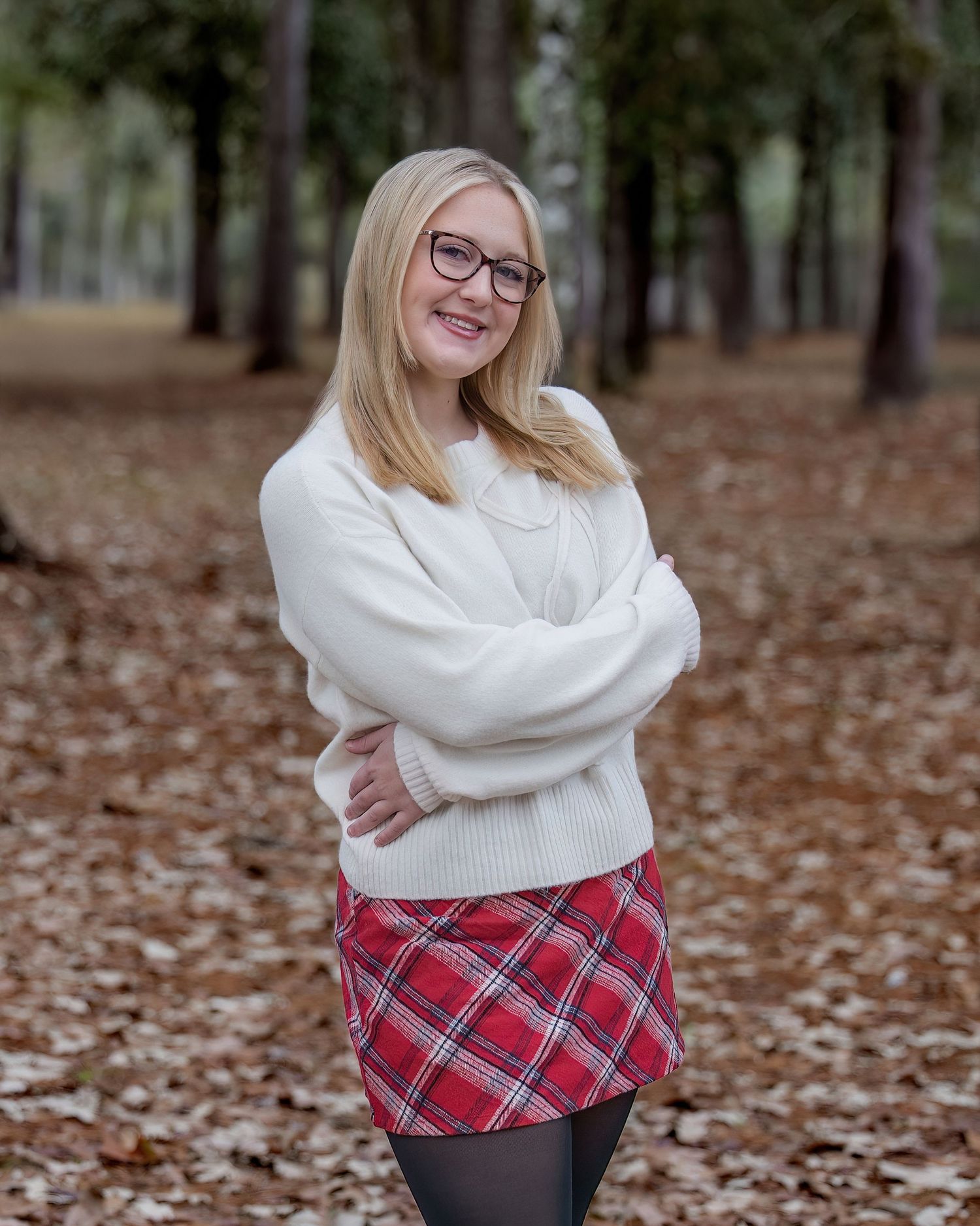 Person wearing a white sweater and plaid skirt poses in a forest covered with fallen leaves.
