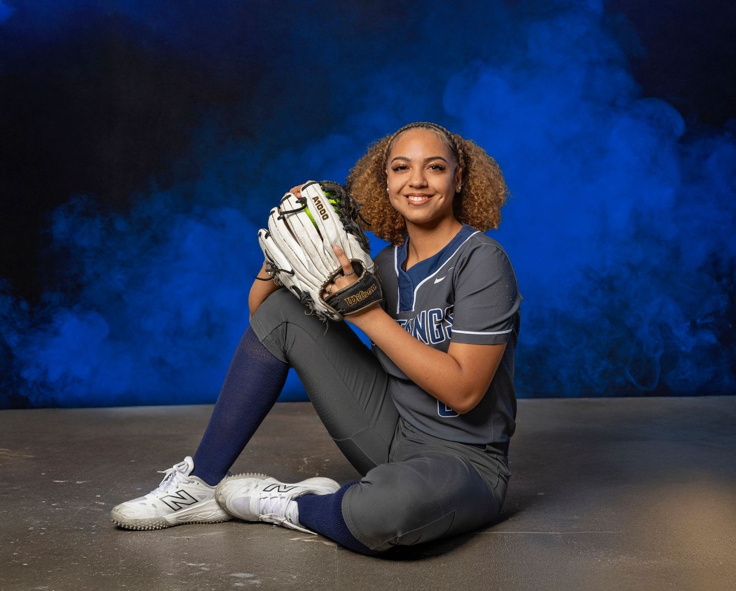 Softball player in grey uniform sits with glove against dramatic blue smoke background for sports portrait.