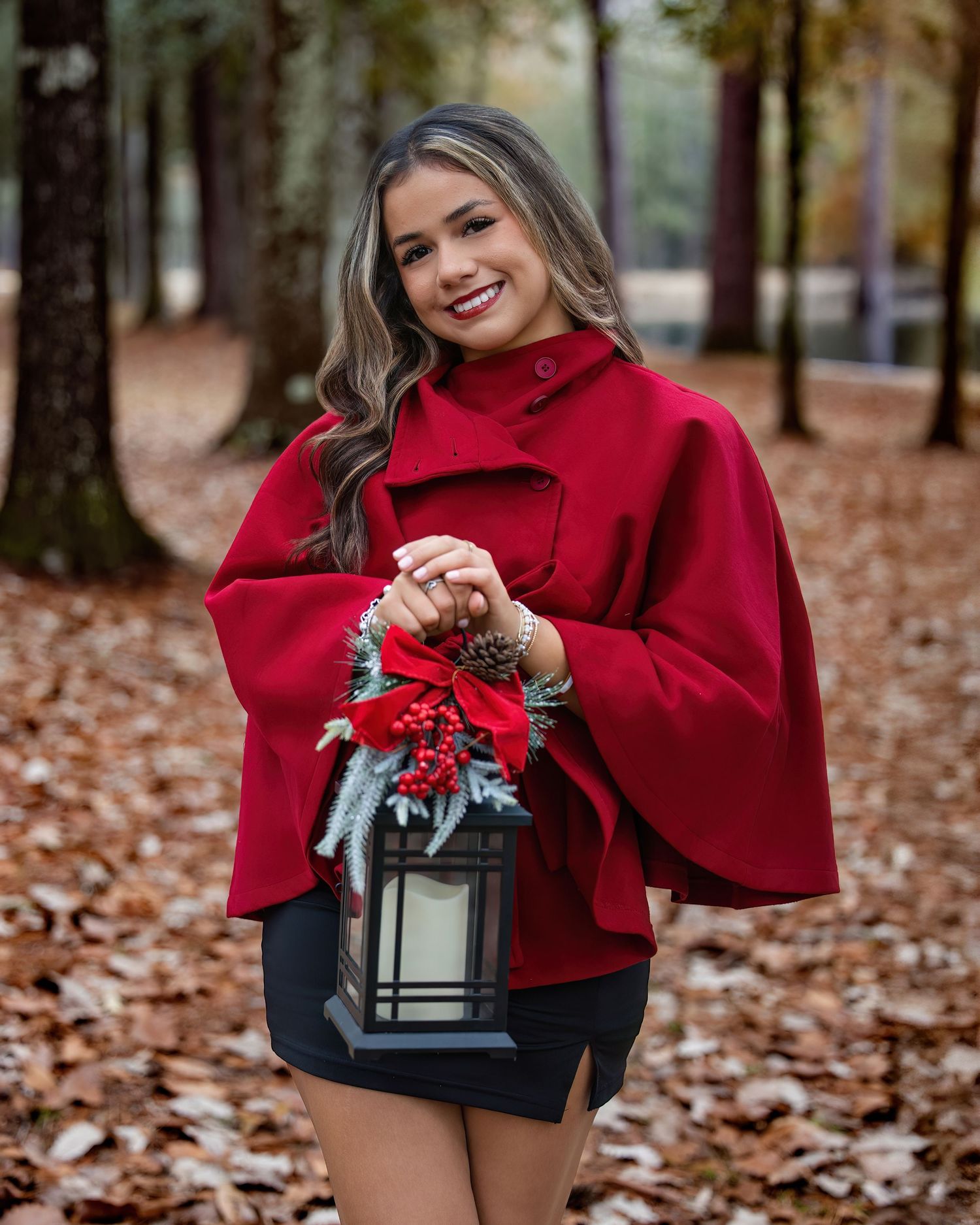 A person in a red sweater poses with a decorative lantern on fallen autumn leaves in an outdoor setting.