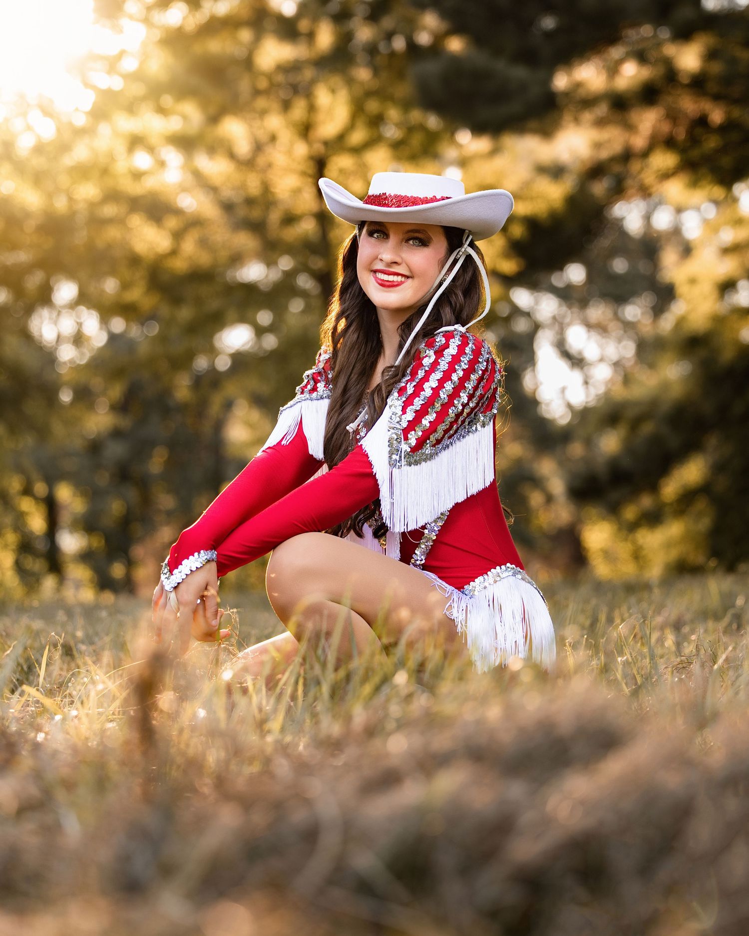 Young woman in red and white cowgirl outfit with fringe and white hat poses in golden sunlit field.