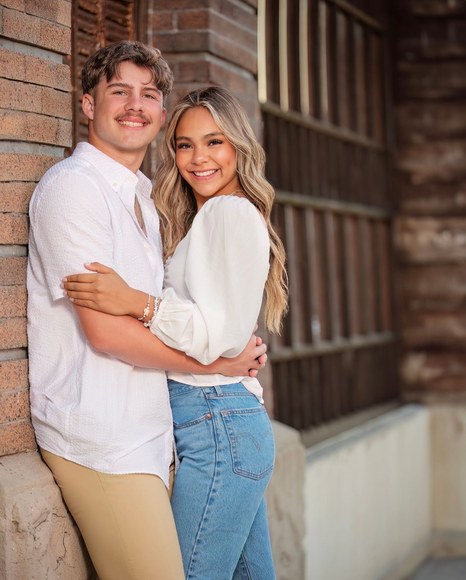 A couple in casual outfits embracing and smiling against a rustic brick wall backdrop.