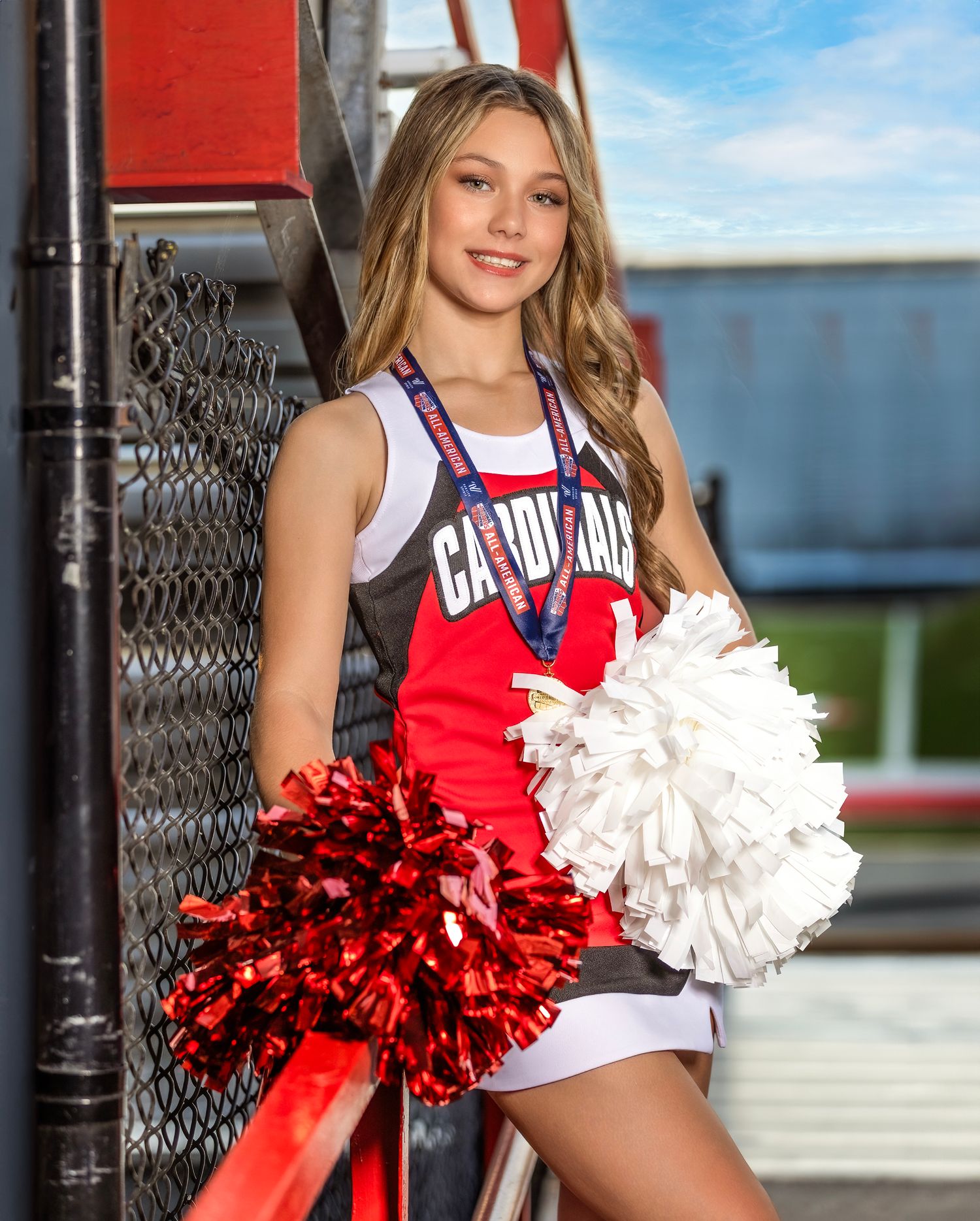 High school cheerleader in red and white uniform poses with pom poms against chain link fence outdoors.