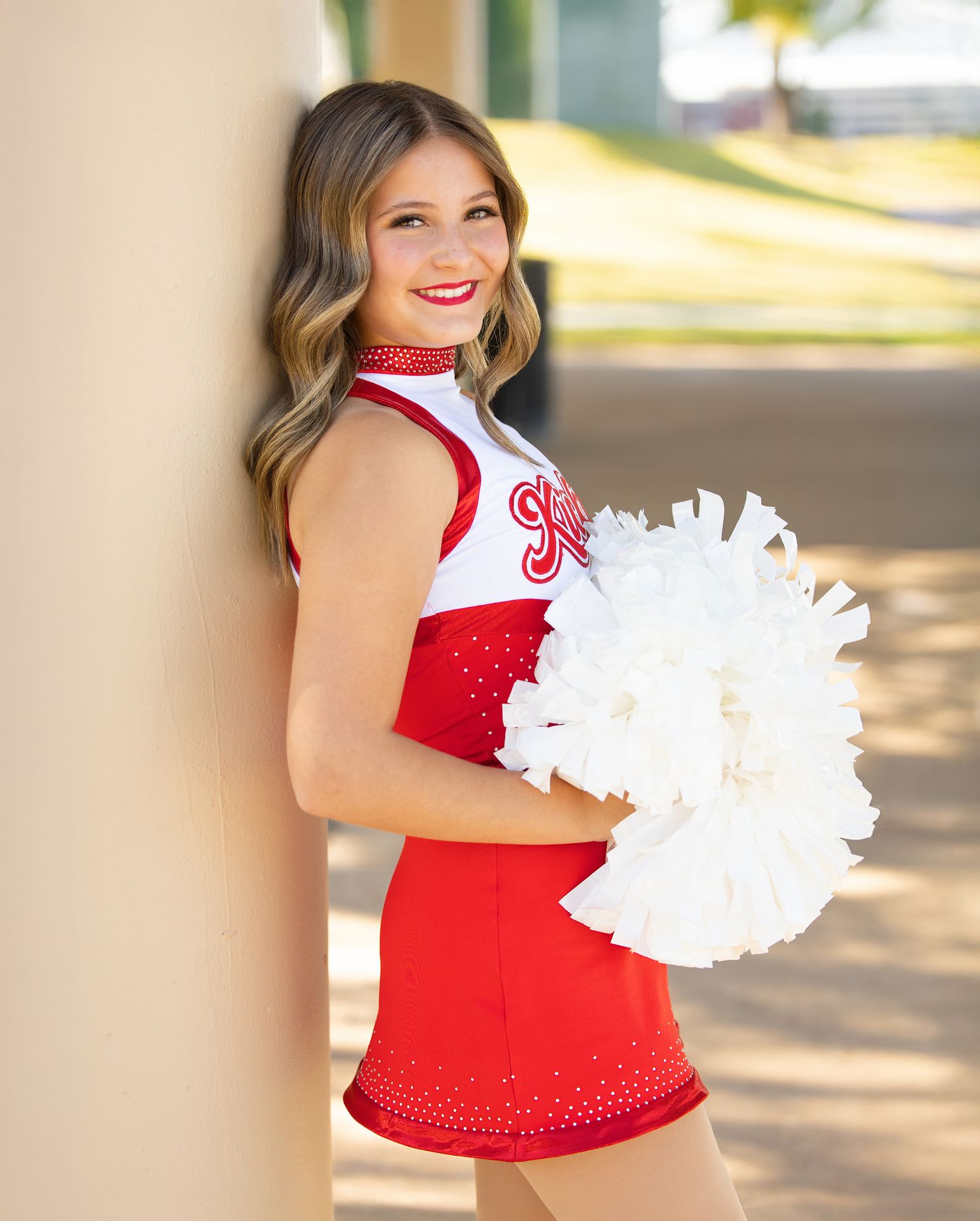 A cheerleader in red and white uniform poses with white pom poms during an outdoor photoshoot.