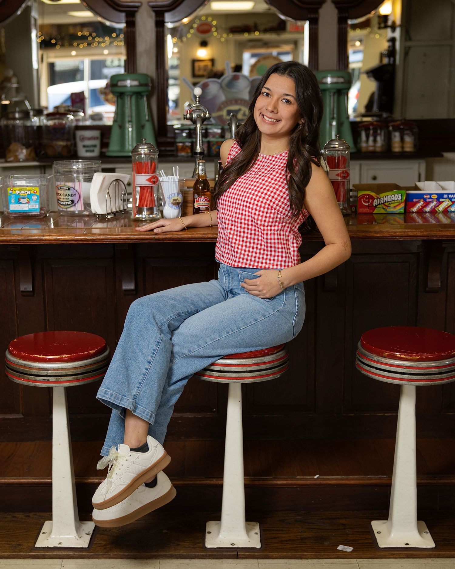 Person in red gingham top and jeans sits casually at vintage diner counter stools.