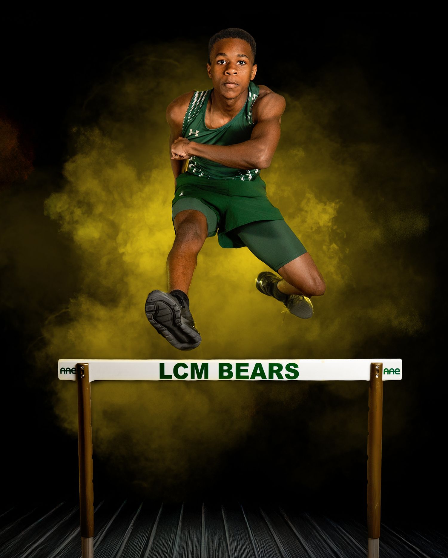 Track athlete in green uniform leaps over hurdle against dramatic yellow smoke background during competition.