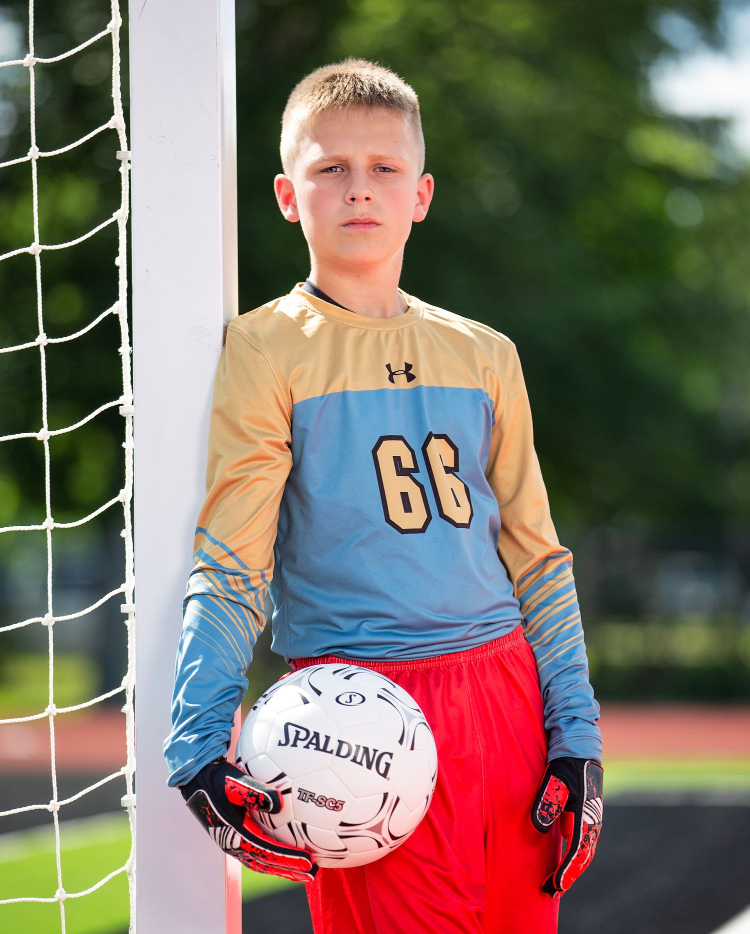 Young soccer goalkeeper in yellow and blue jersey with red shorts stands ready at goal post with ball in hand.