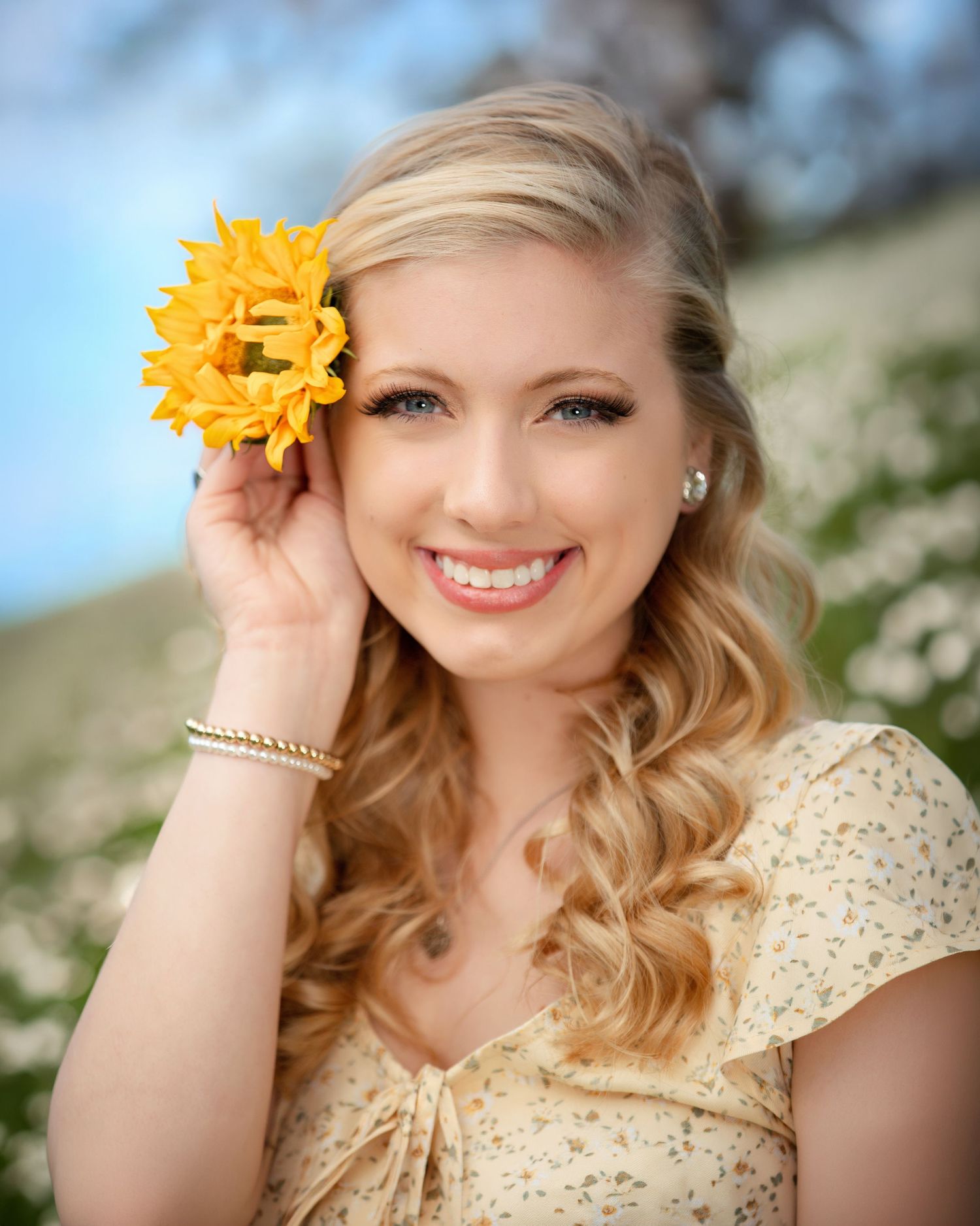 Smiling blonde woman holding a yellow sunflower in her hair, wearing a floral dress in a field of white flowers.