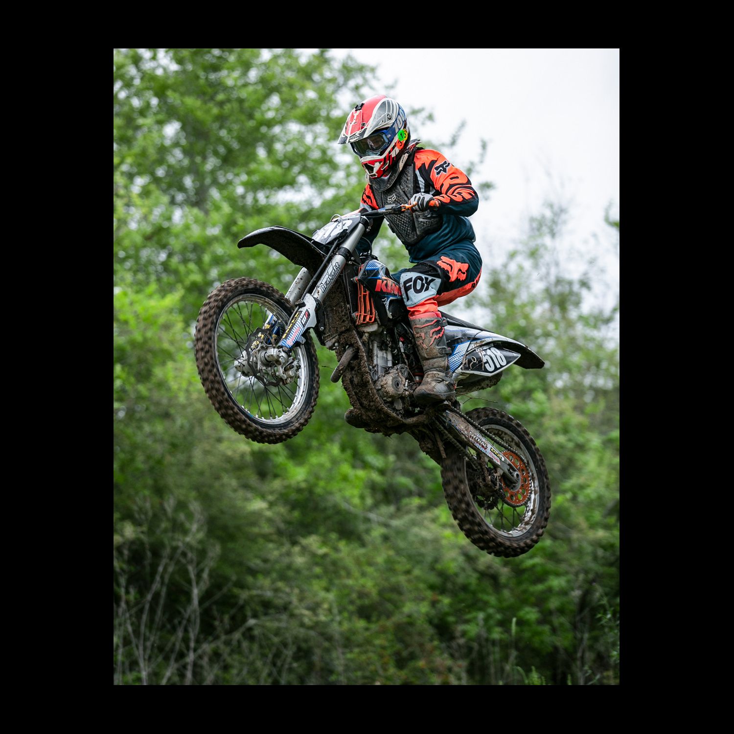 Motocross rider in orange gear catches air on a dirt bike during an outdoor race with trees in background.