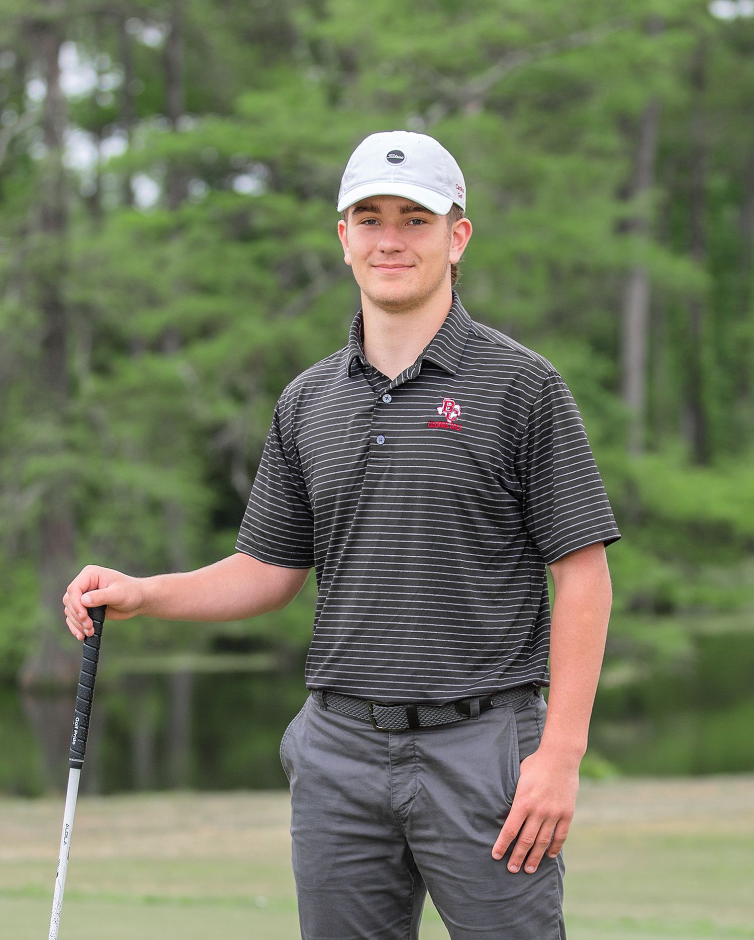 Golfer in striped polo and gray pants poses with club on green course surrounded by pine trees.