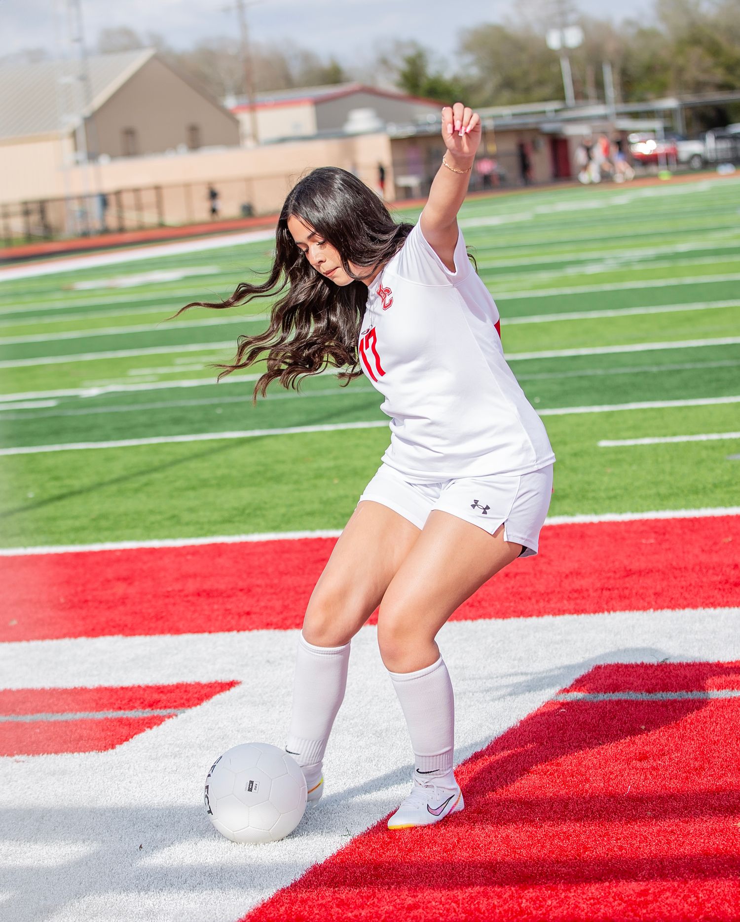 Soccer player in white uniform dribbles ball on red turf field during daytime practice session.