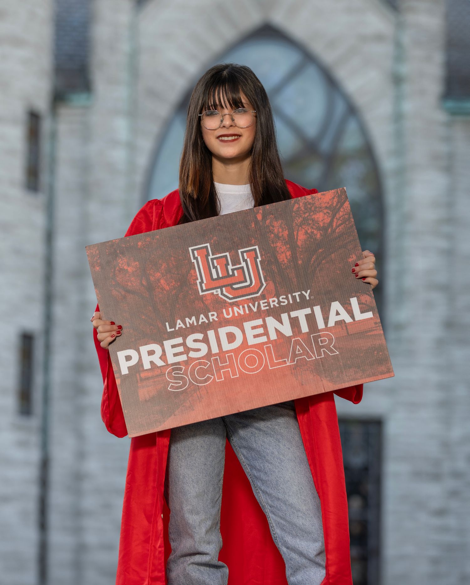 Student in red graduation gown holds up Lamar University Presidential Scholar sign during outdoor photo session.