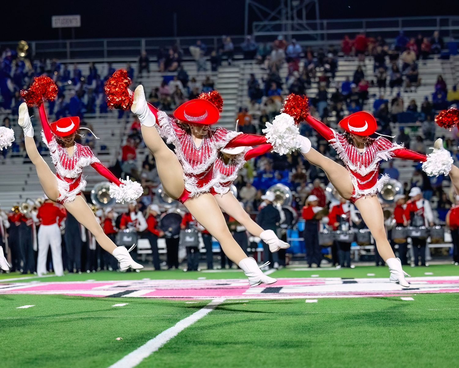 Cardinal Strutters performing synchronized split jump on football field in red and white uniforms.