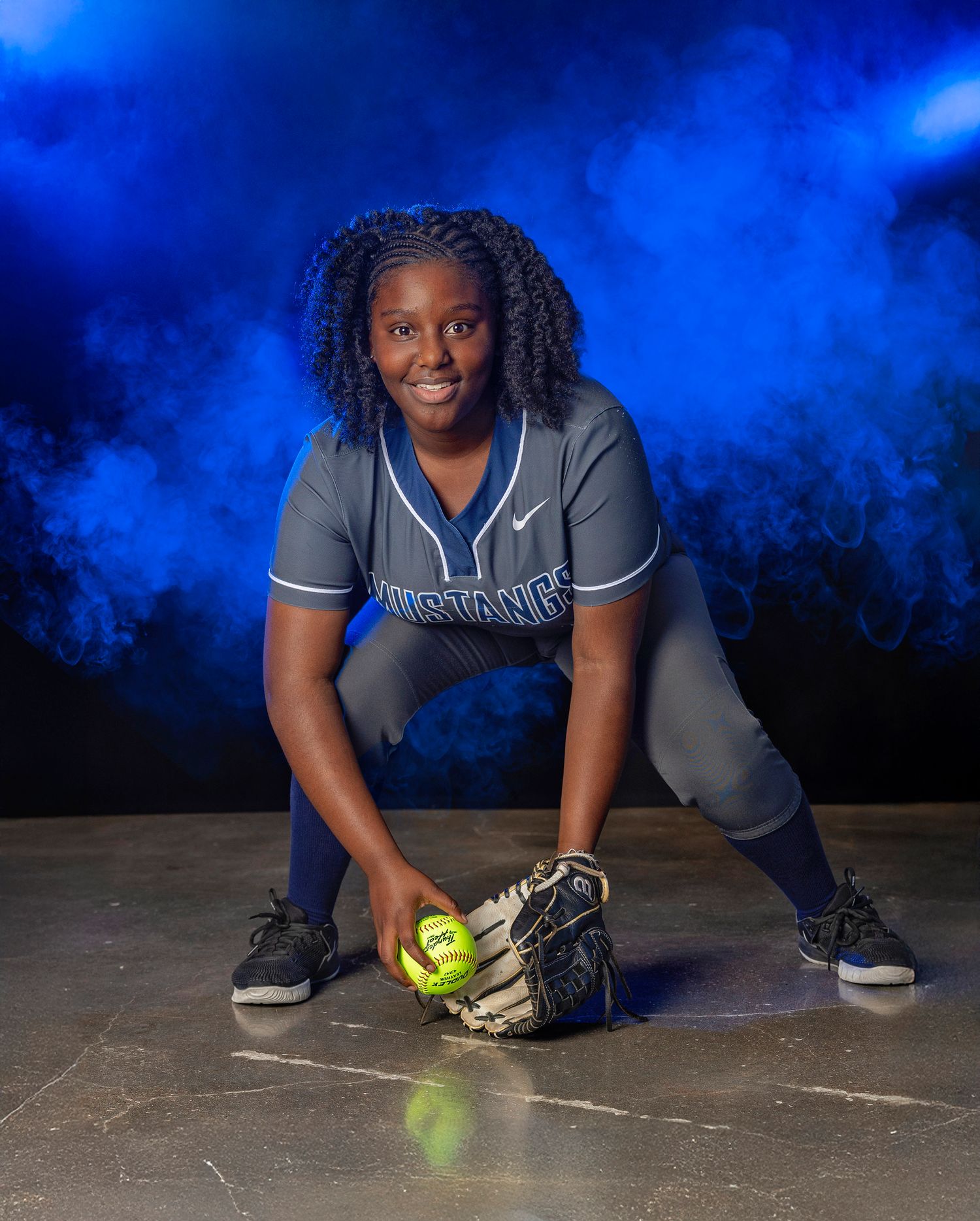 A softball player in a gray uniform poses with a glove and ball against a dramatic blue background.