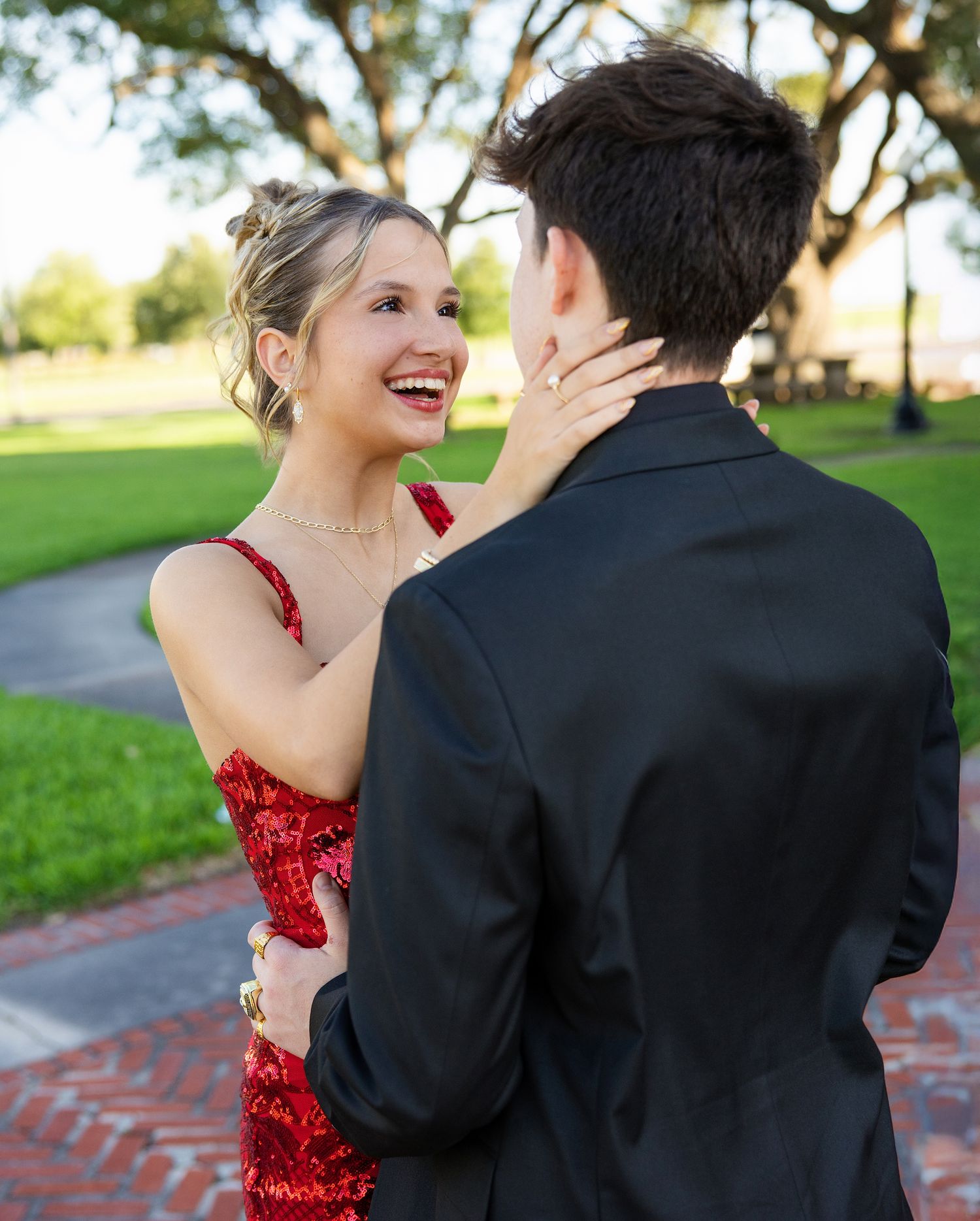A couple shares a joyful moment in formal attire, with a red sequined dress and black suit against a green outdoor backdrop.