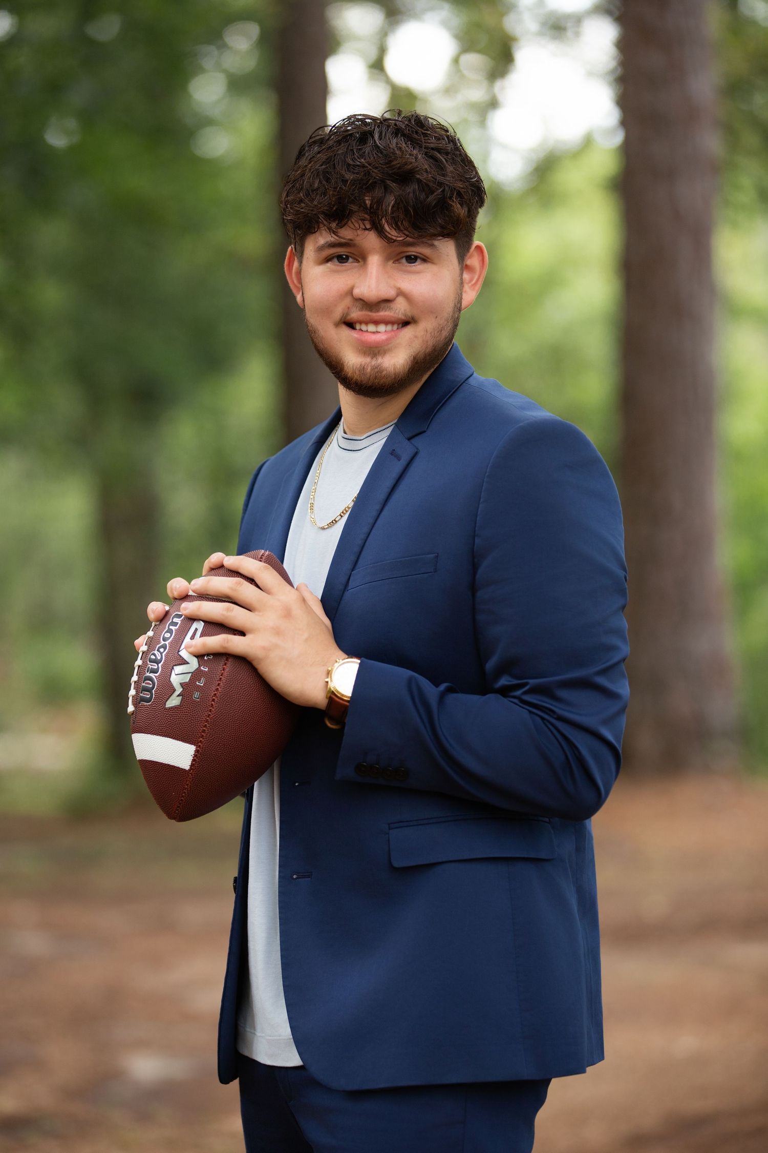 Senior Football player in a navy blue blazer and white shirt holds a football while posing in an outdoor wooded setting.