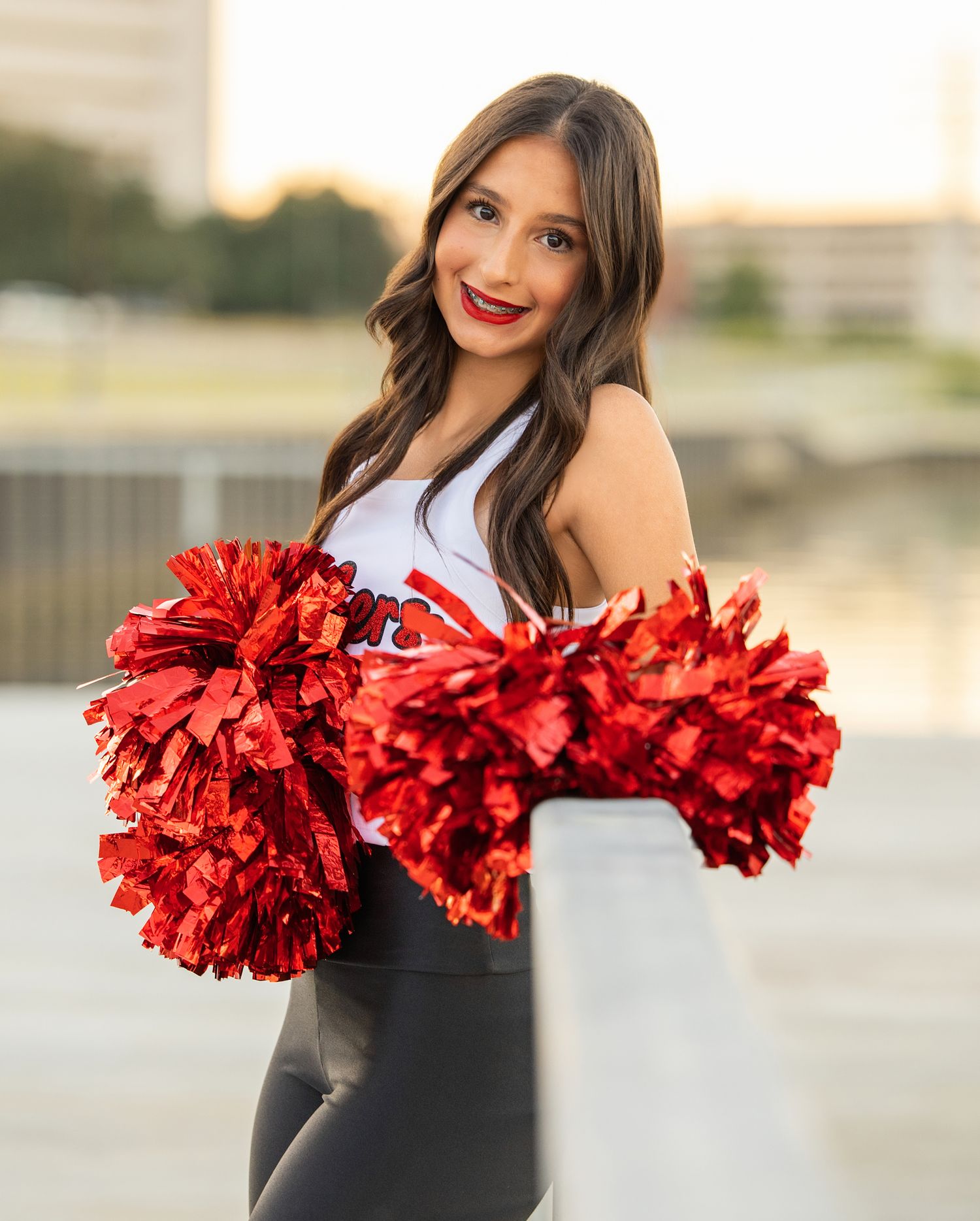 A cheerleader in a black outfit holds red and metallic pom poms while posing outdoors at sunset.