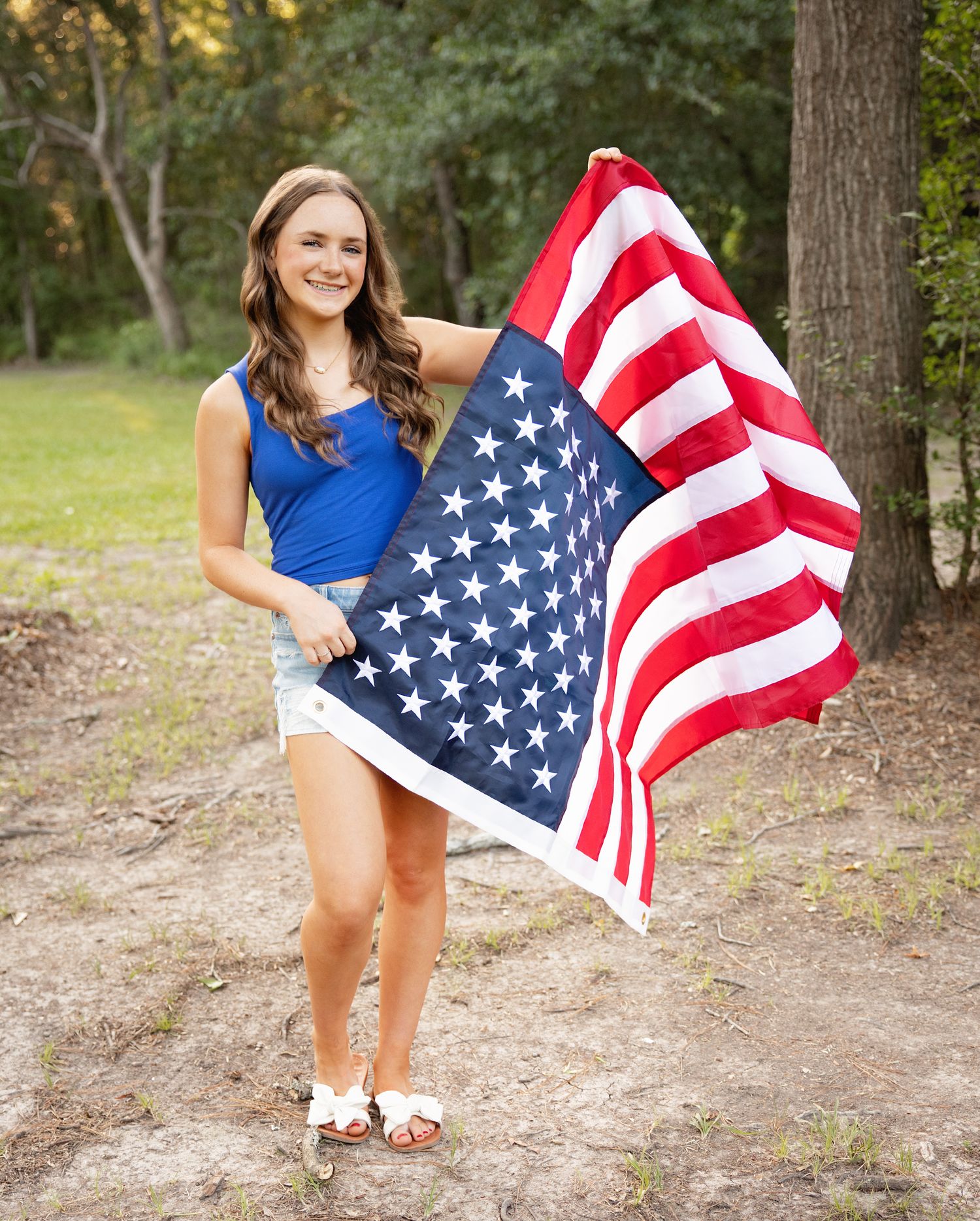 A person in a blue tank top and white shorts holds an American flag outdoors in a wooded setting.