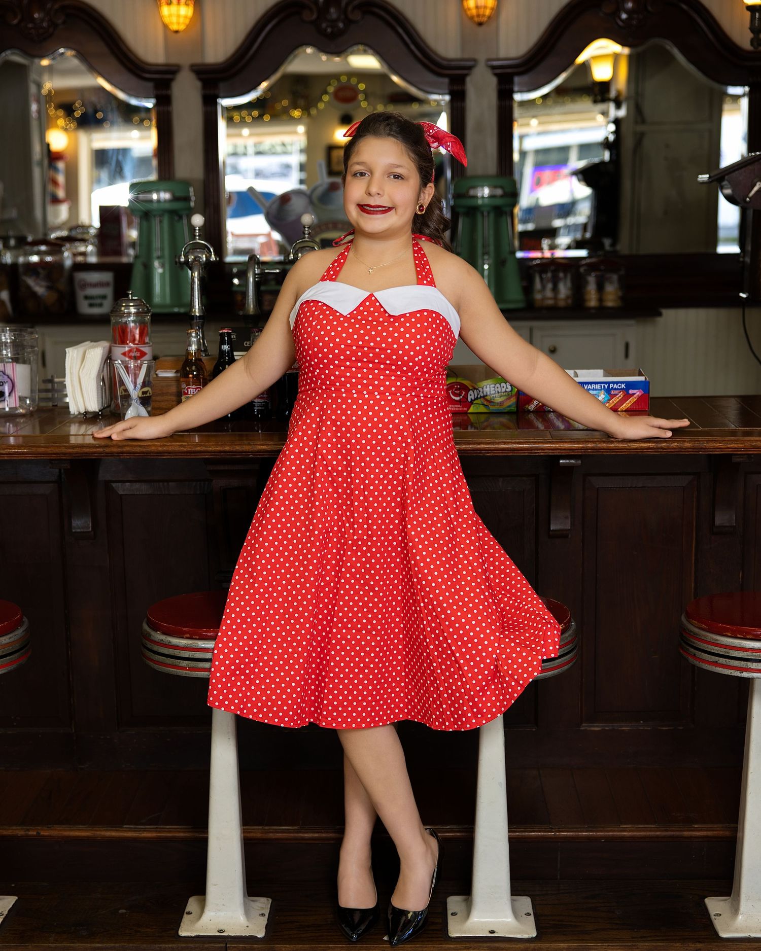 Someone in a red and white polka dot dress poses at a vintage diner counter with bar stools.