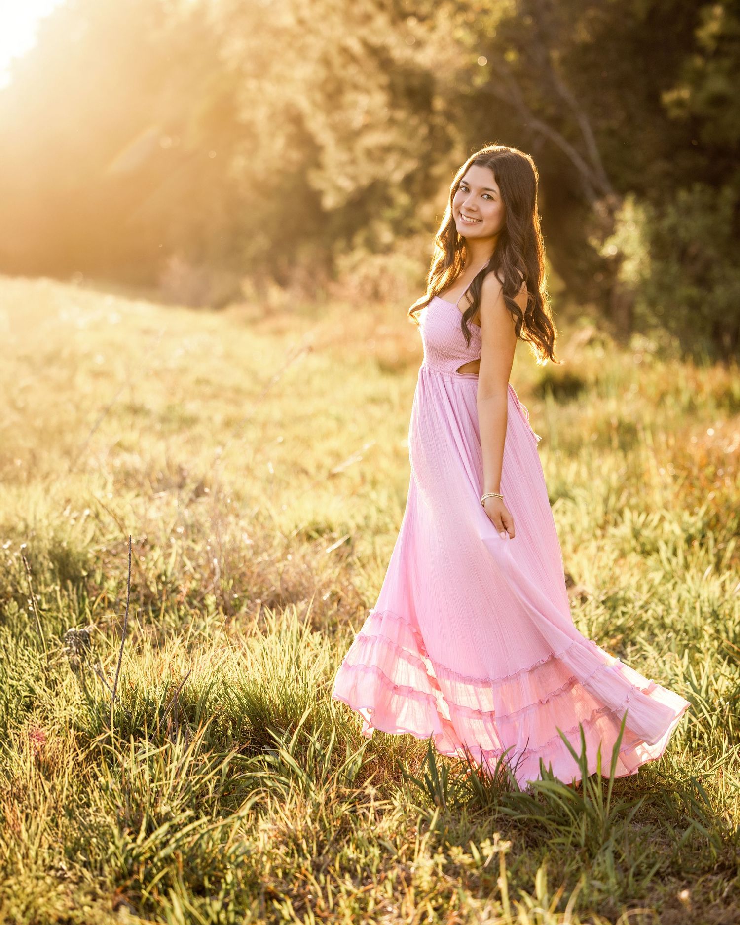 Young woman in flowing pink maxi dress twirling in sunlit meadow during golden hour outdoor portrait photography.