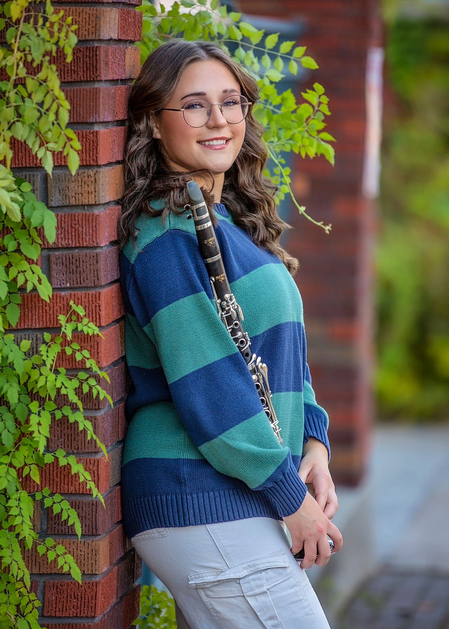 A smiling teenage girl with glasses holds a clarinet, wearing a blue and green striped sweater while leaning against a brick wall.