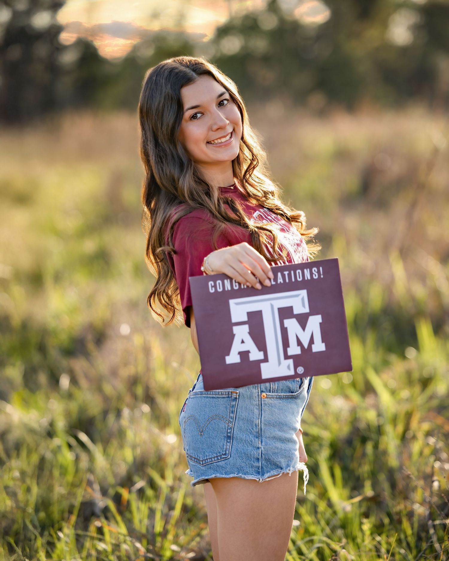 Smiling young woman holding a Texas A&M Congratulations sign in a sunlit field, wearing a maroon top and denim skirt.