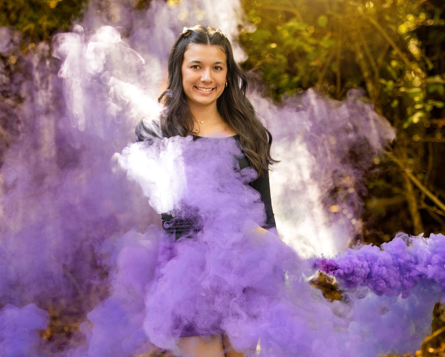 A person poses outdoors surrounded by vibrant purple smoke bombs creating a dreamy atmospheric effect.