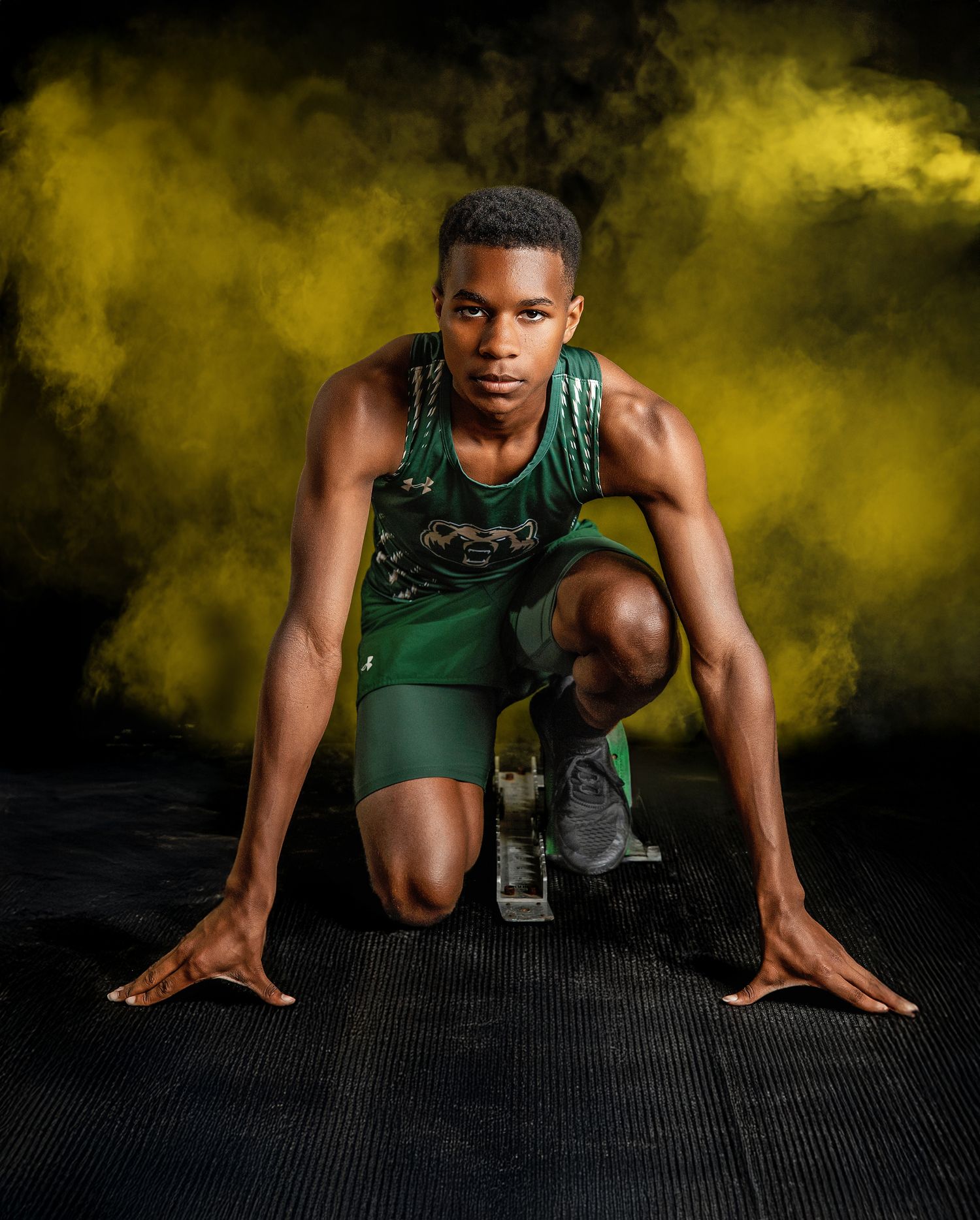Athletic runner in green uniform crouches in starting position against dramatic yellow smoky background.