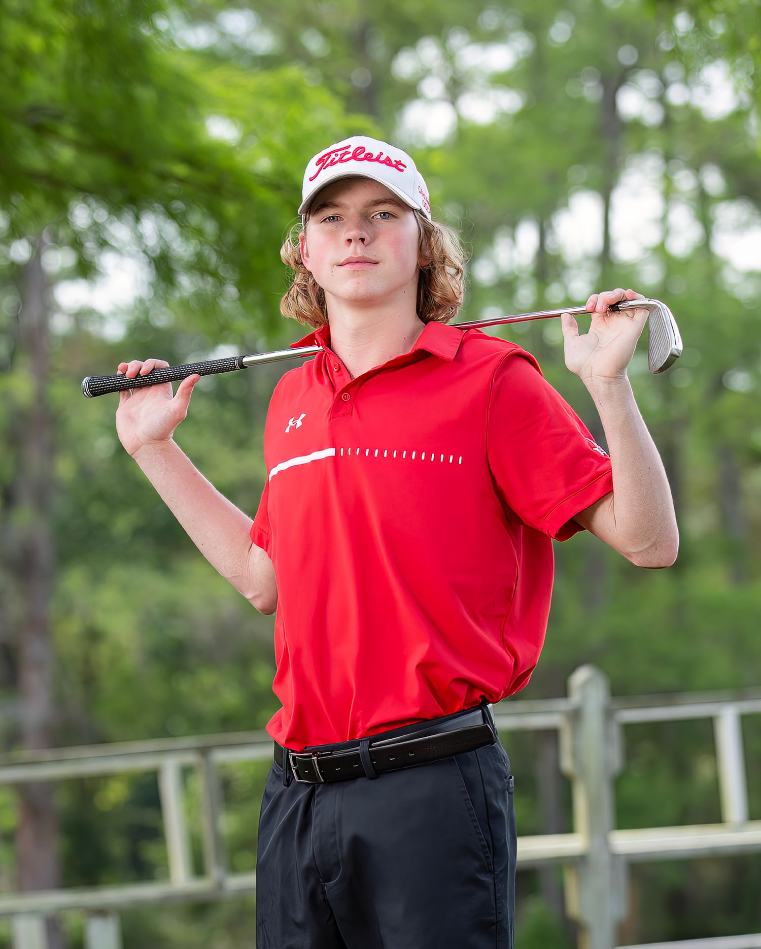 Young golfer in red polo shirt and black pants follows through on swing at outdoor golf course.
