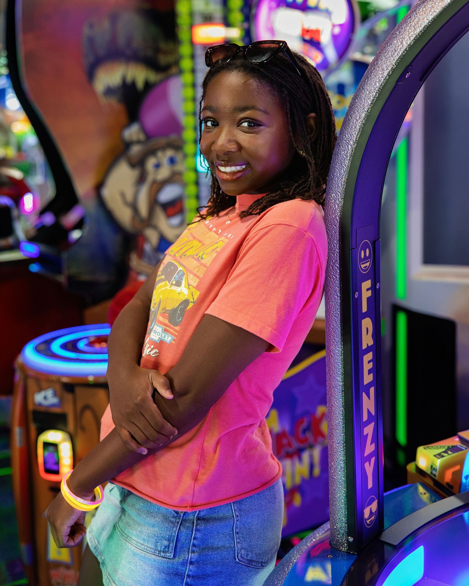 Young woman in pink t-shirt smiling beside a Frenzy arcade game in a colorful arcade.
