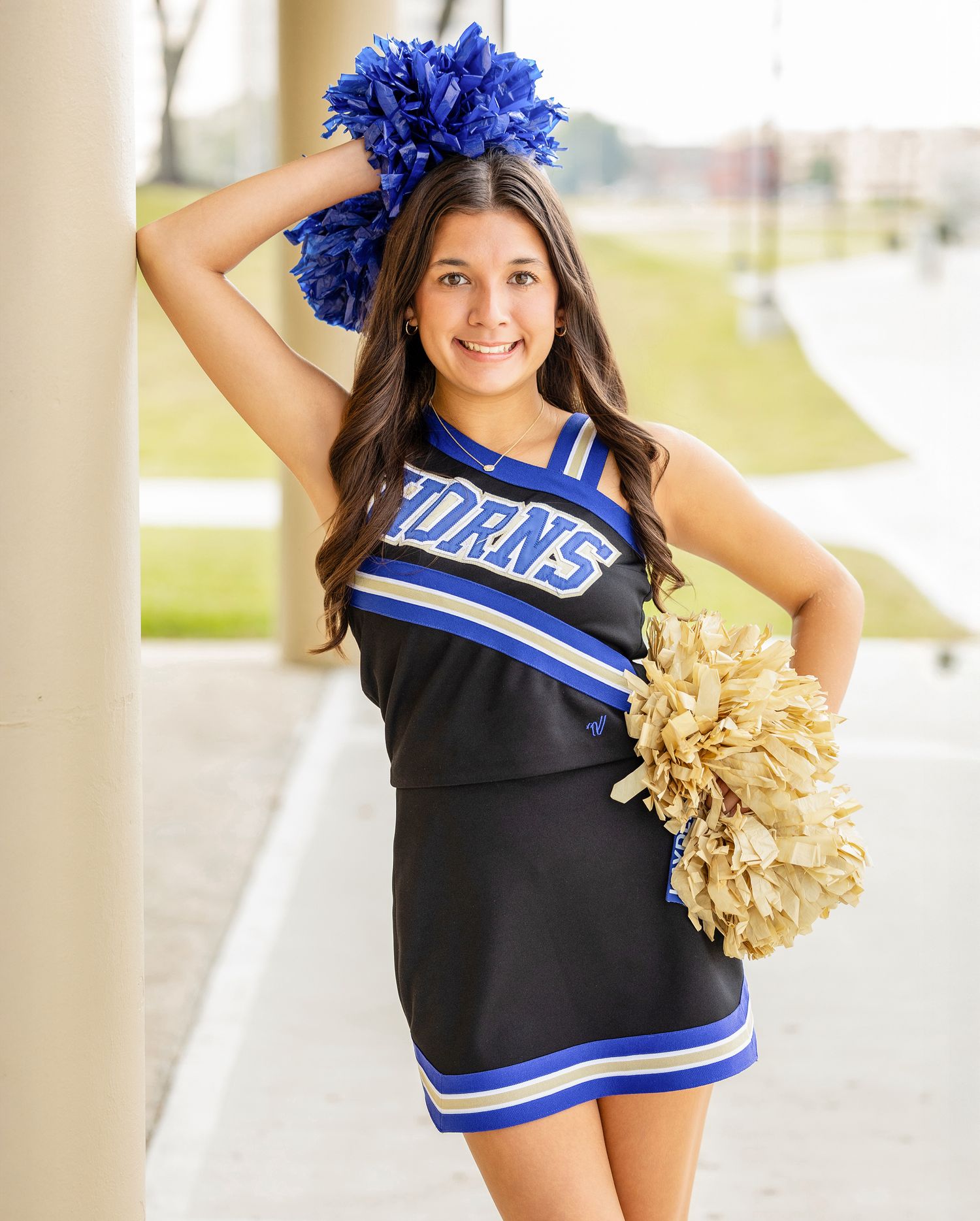 A cheerleader in blue and black uniform with pom poms performs energetic dance moves in a covered outdoor area.