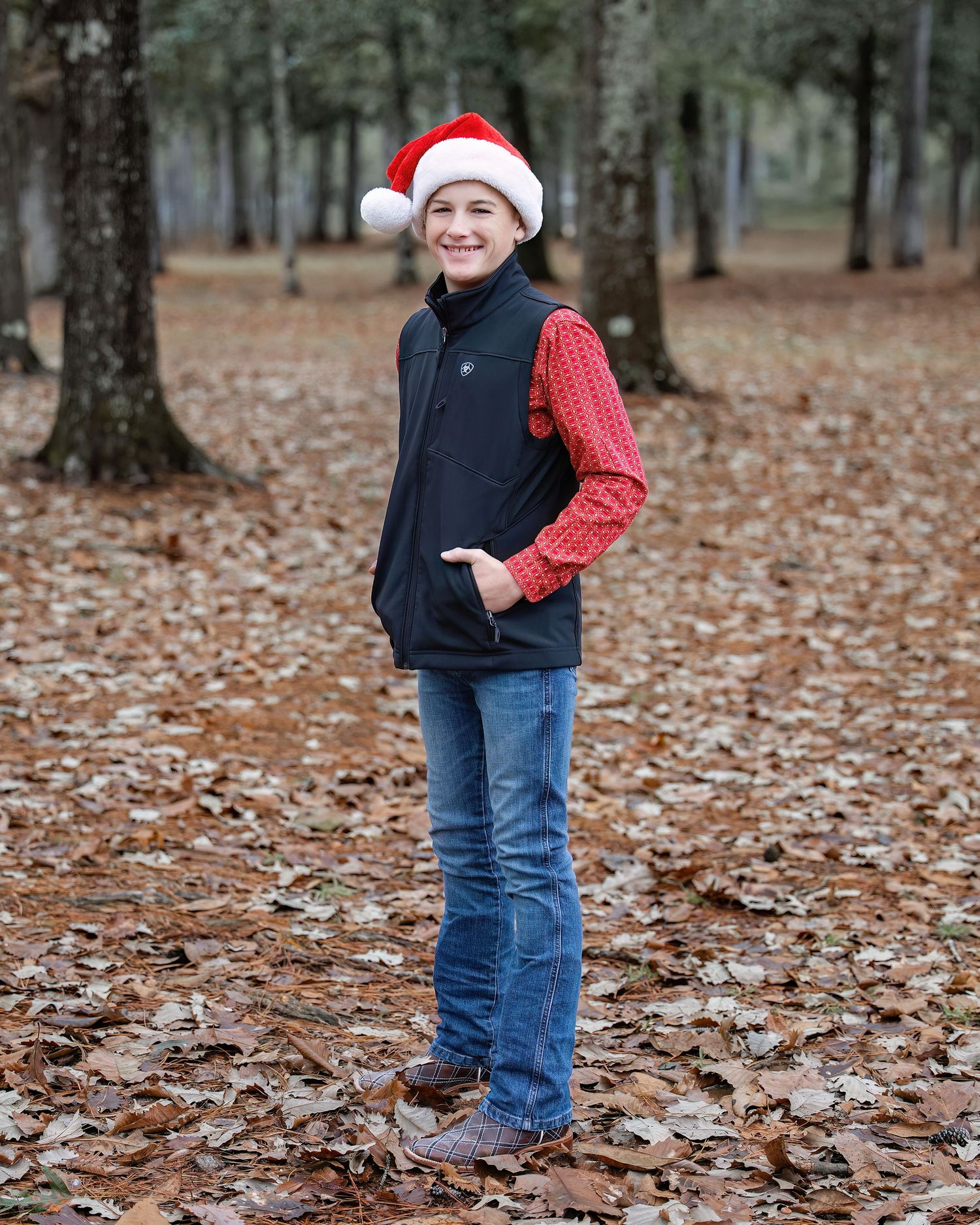 Child wearing Santa hat and winter clothes plays in fallen leaves during holiday photoshoot.