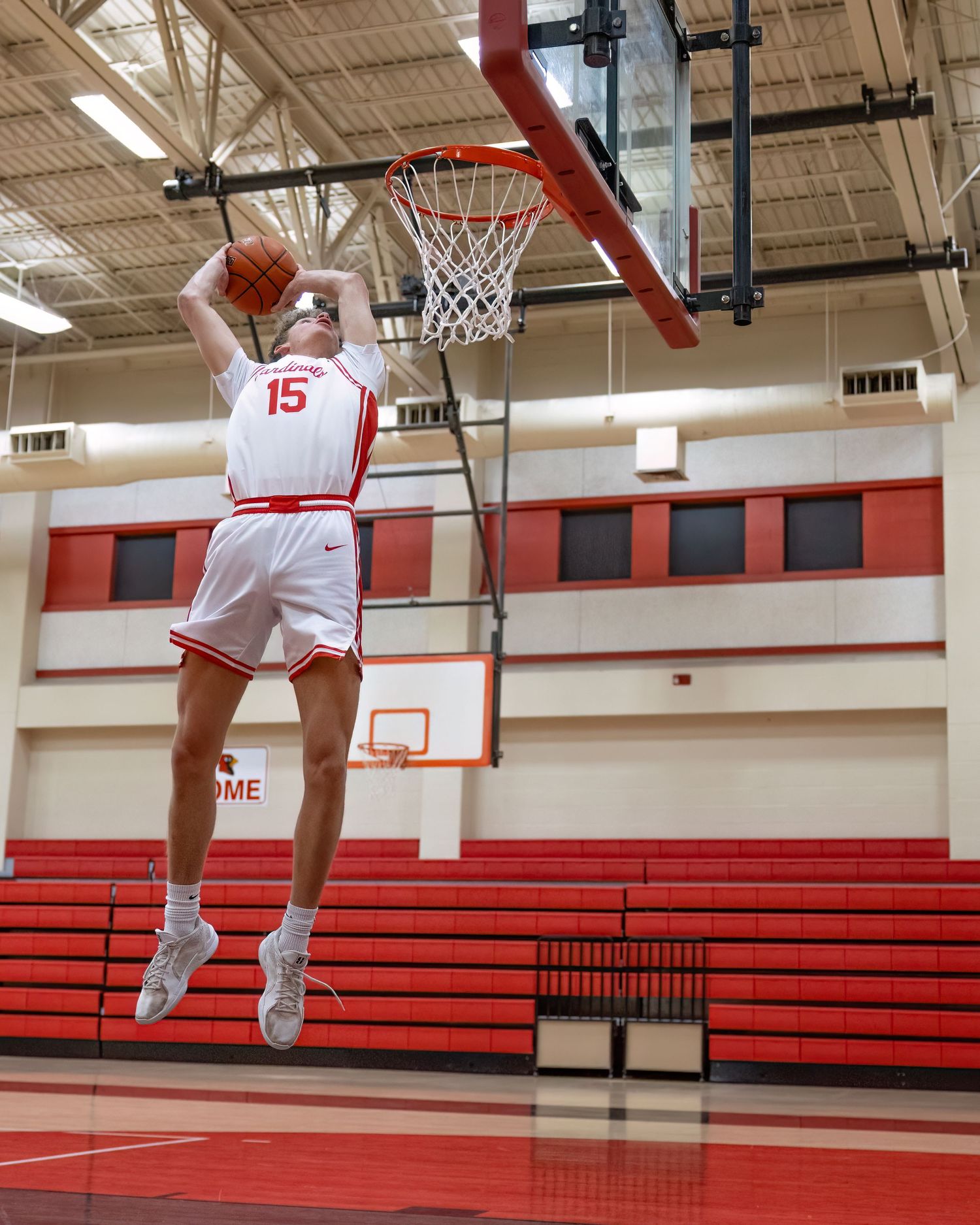 Basketball player in white jersey performs a slam dunk sequence in an empty gymnasium with red bleachers.