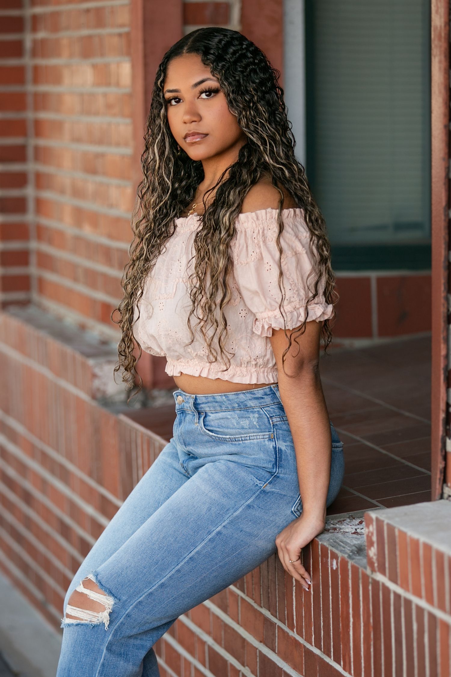 High School Senior with curly hair poses in ripped blue jeans and white crop top against brick wall backdrop.