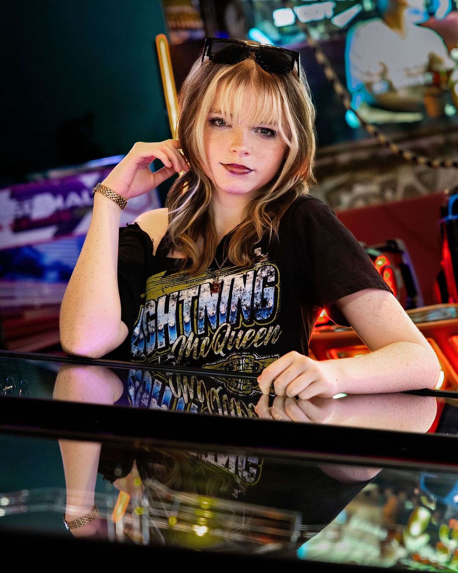 Young woman in Lightning McQueen shirt poses at arcade pinball machine with sunglasses on head.