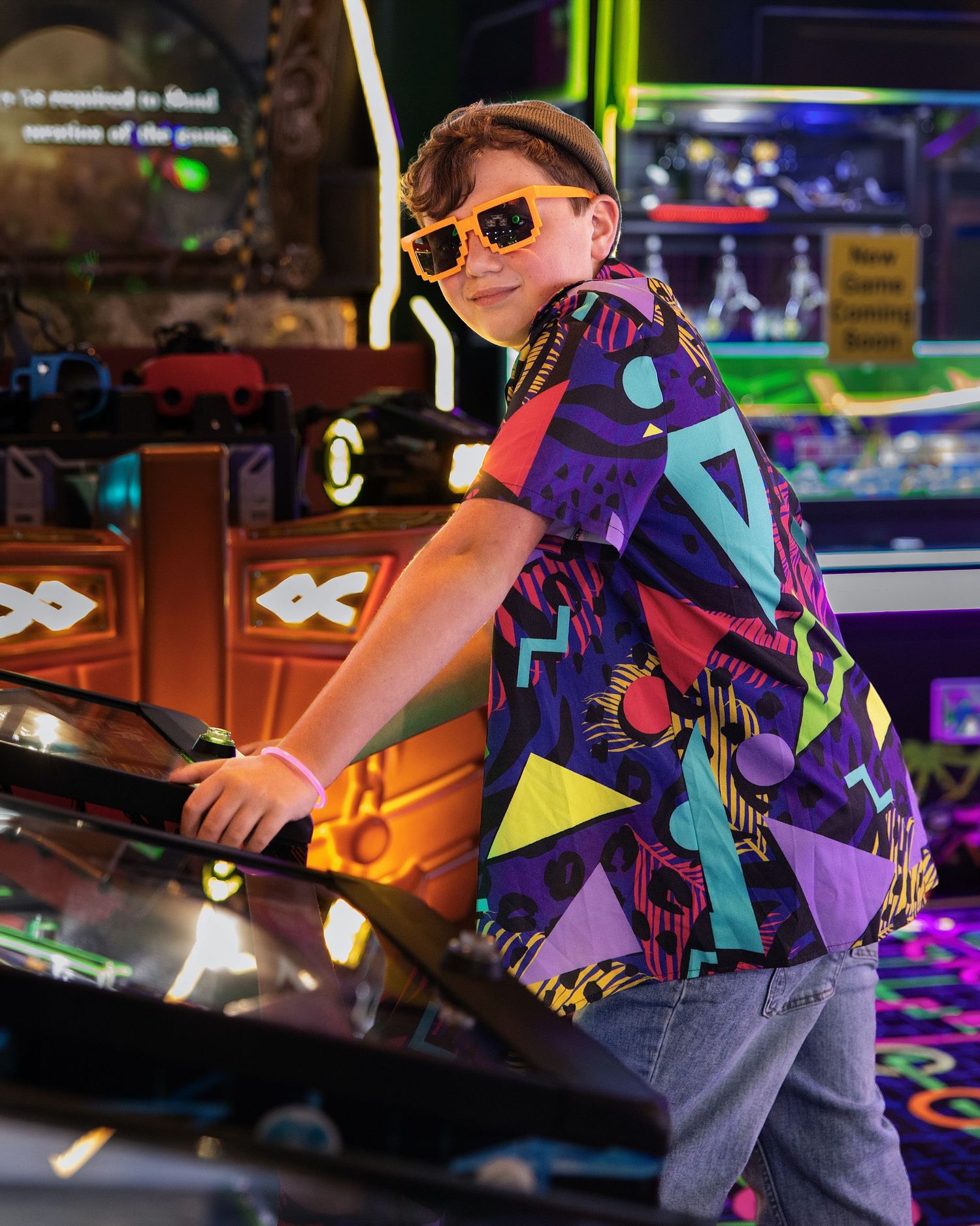 Boy in colorful 80s-style shirt and orange sunglasses playing arcade games in a neon-lit game room.
