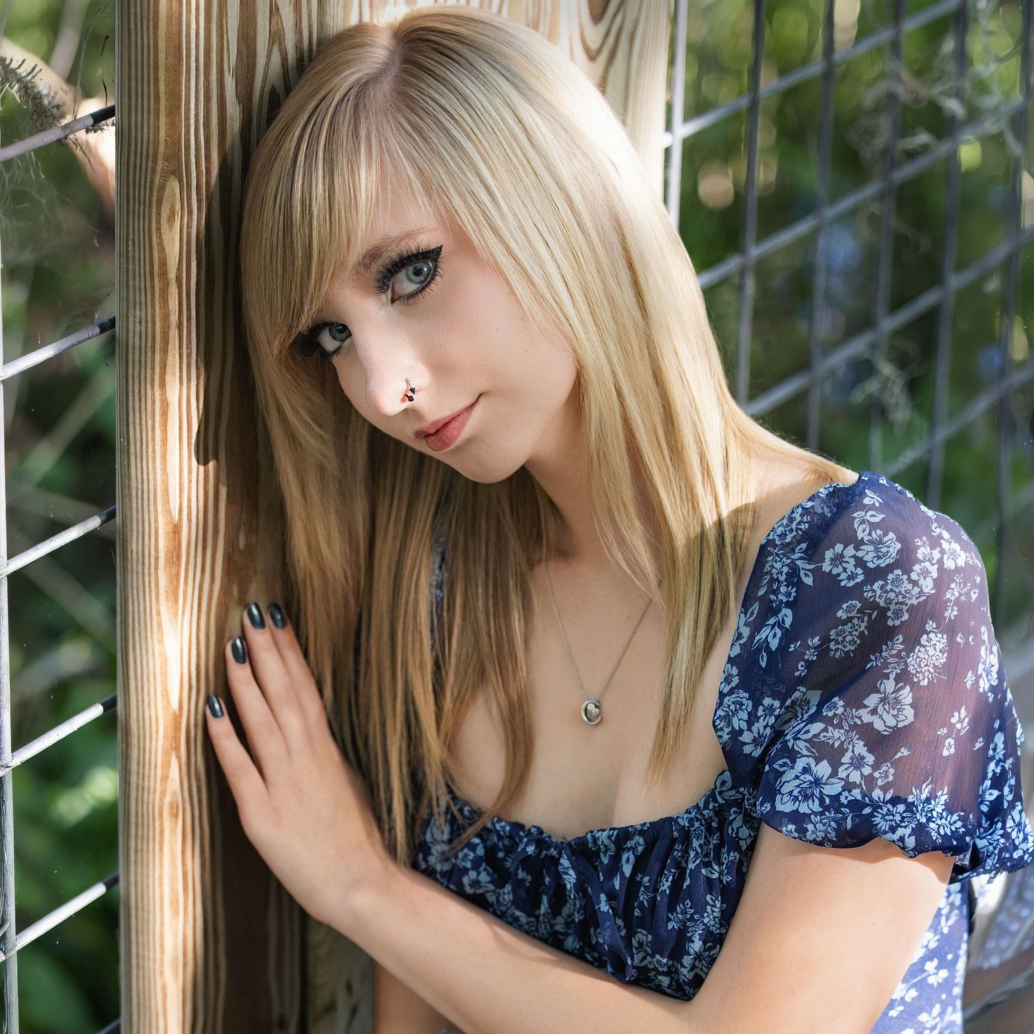 Young blonde woman in floral blue dress leaning against wooden post outdoors, wearing necklace and nose ring.