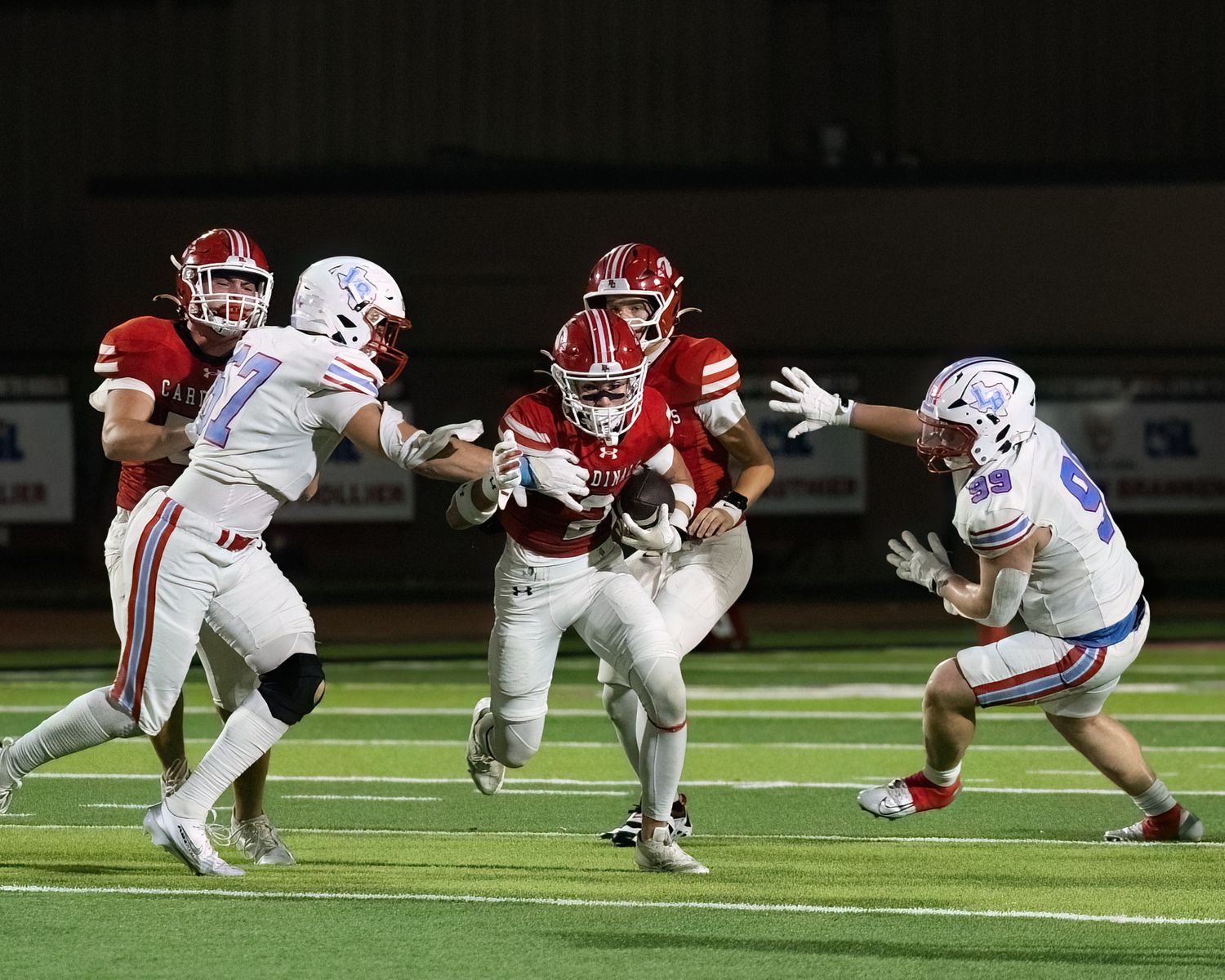 Football players collide during a tackle attempt as a white-uniformed player evades defenders in red jerseys.