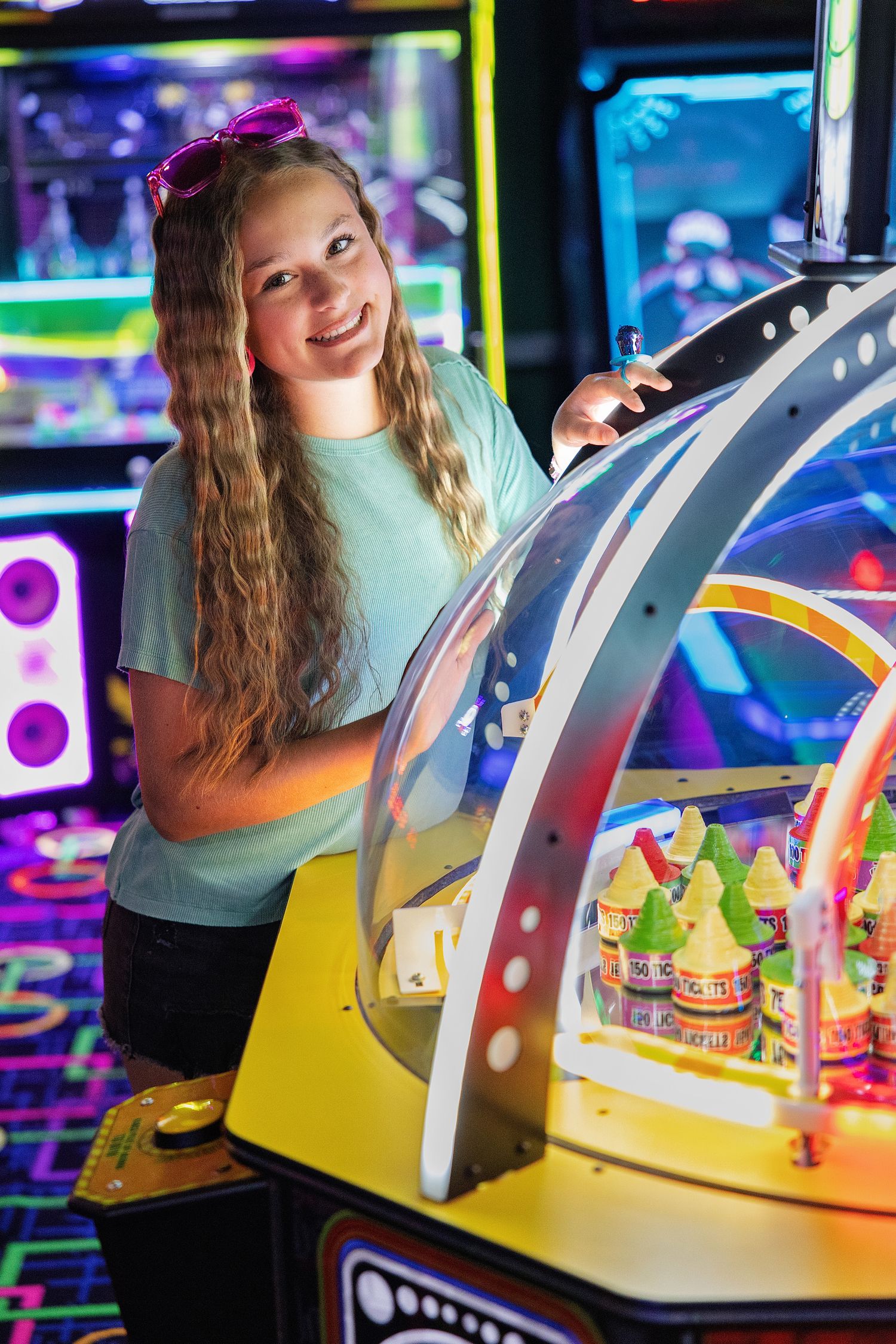 Smiling teenage girl with pink sunglasses playing an arcade game in a colorful neon-lit amusement arcade.