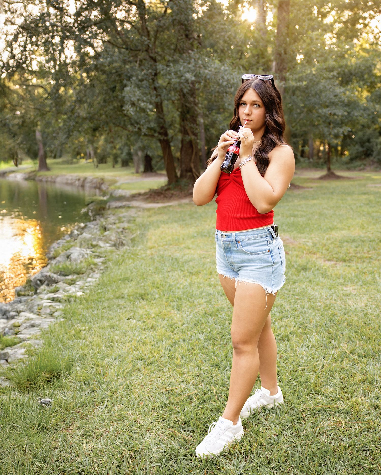 A person in a red tank top and denim shorts poses in a grassy field at sunset with trees in the background.